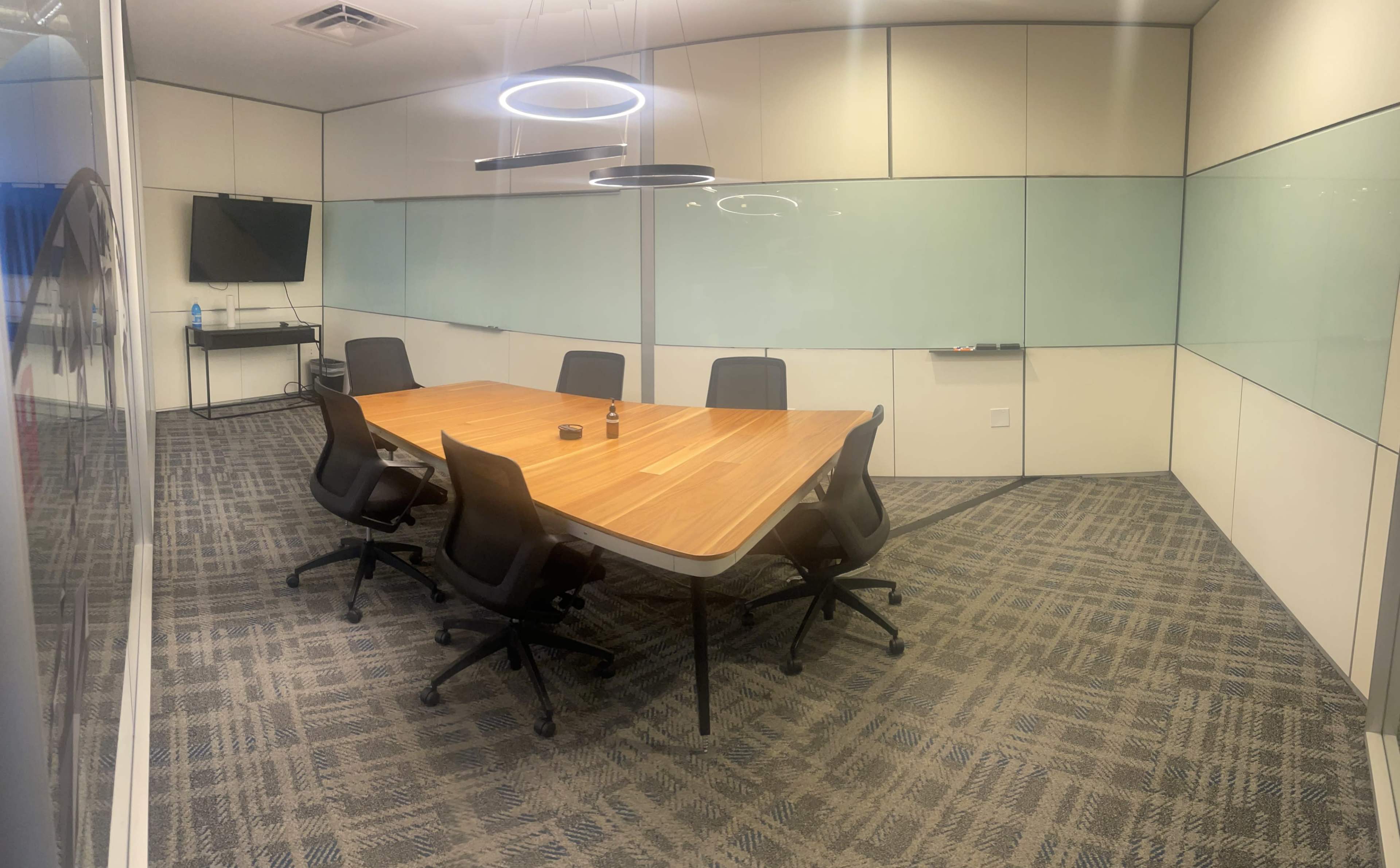 A modern conference room features a large wooden table surrounded by six ergonomic chairs, with a television and glass walls.