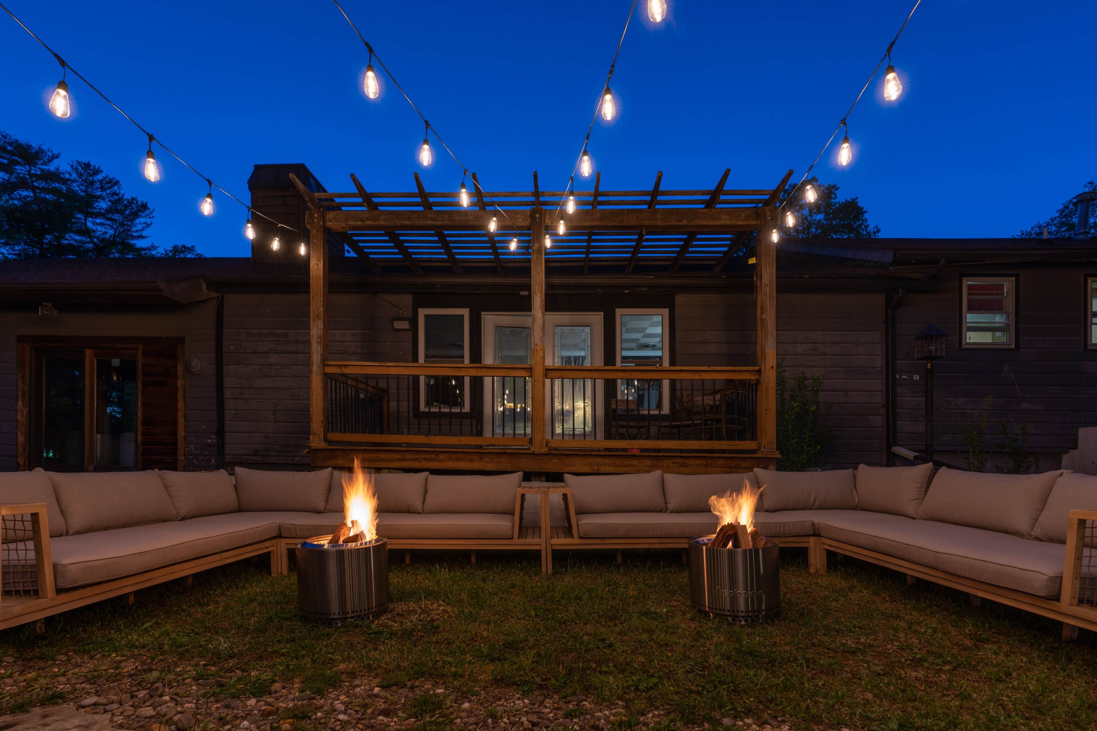 An outdoor seating area with two fire pits on a lawn, surrounded by a large sectional sofa and strung lights overhead at dusk.