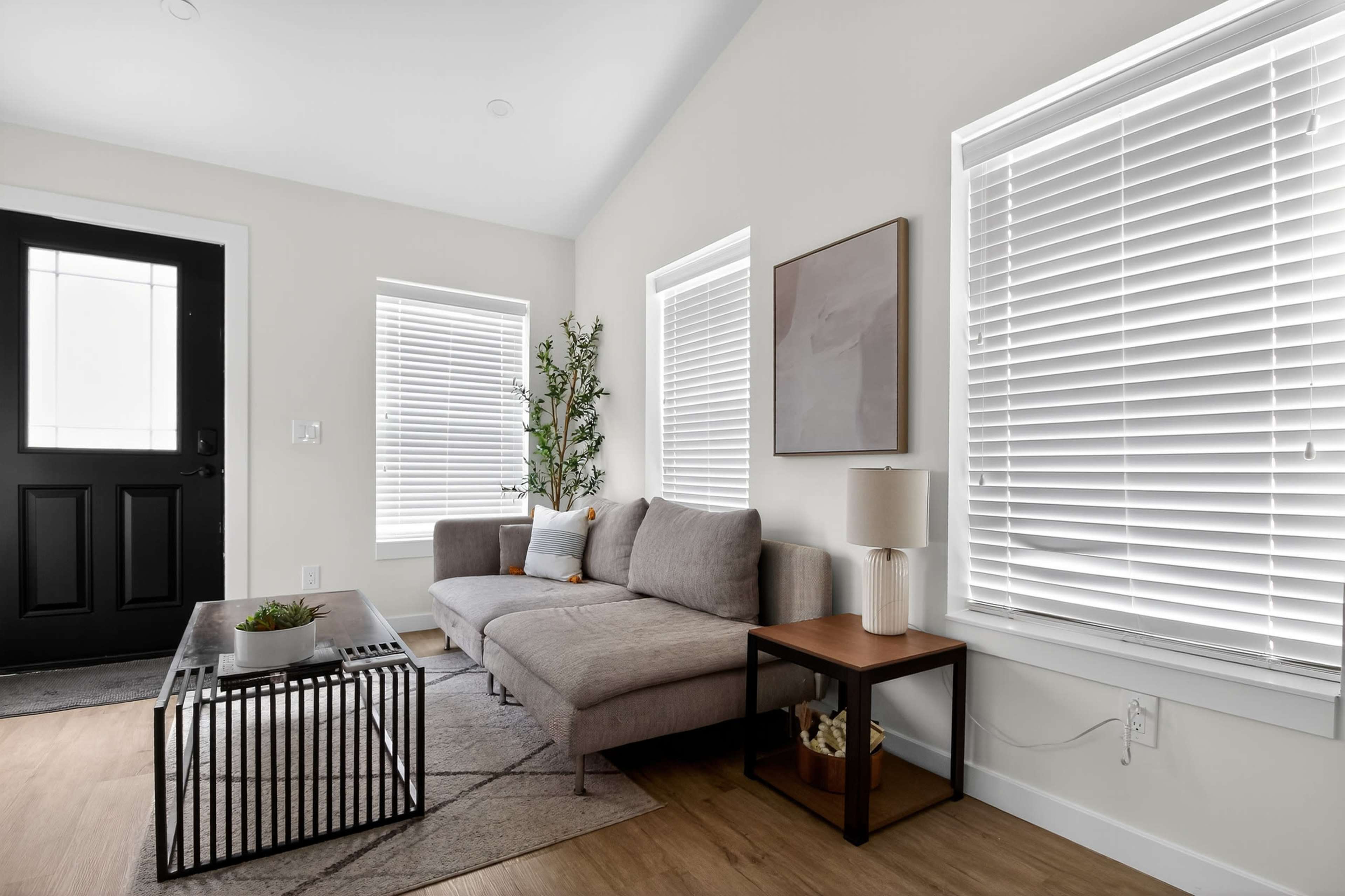 The image depicts a modern living room featuring a gray sofa, a coffee table with a black metal frame, and large windows with white blinds.