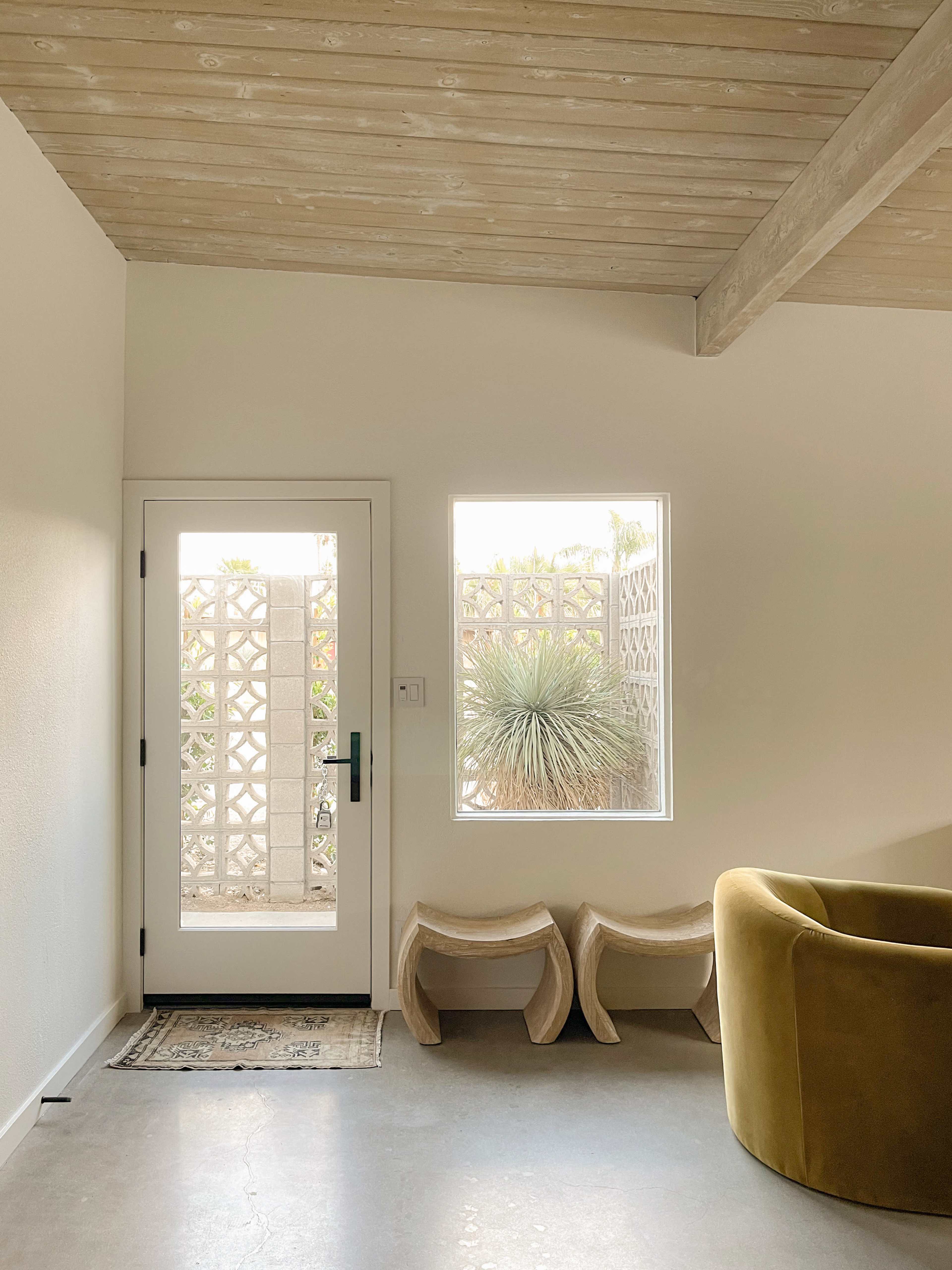 A minimalist interior space featuring a white entrance door with a window beside it, a textured wall, two curved furniture pieces, and a rug on a concrete floor.