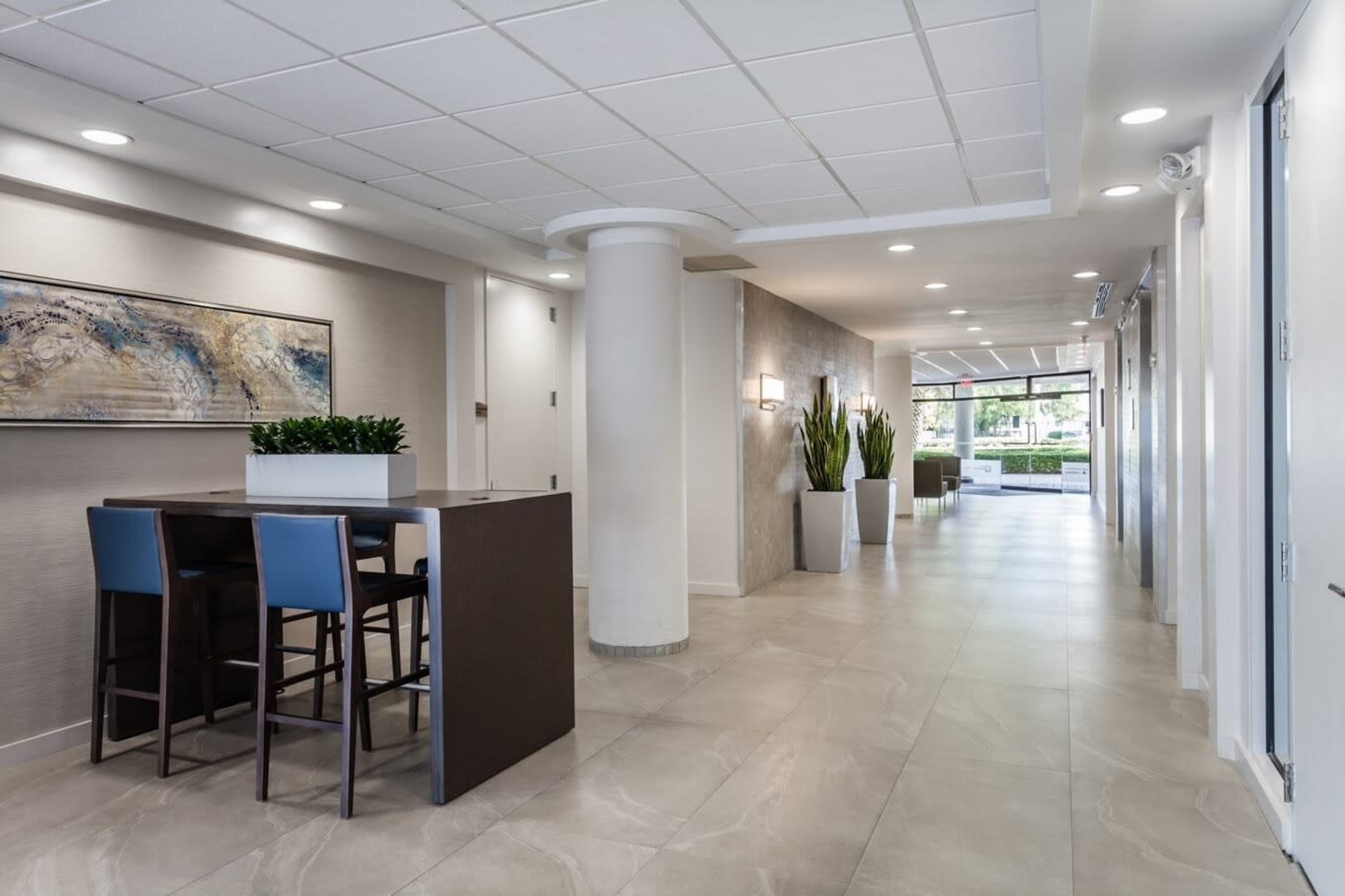 A modern hallway with a small seating area featuring two blue chairs next to a table, leading to large windows with potted plants on either side.