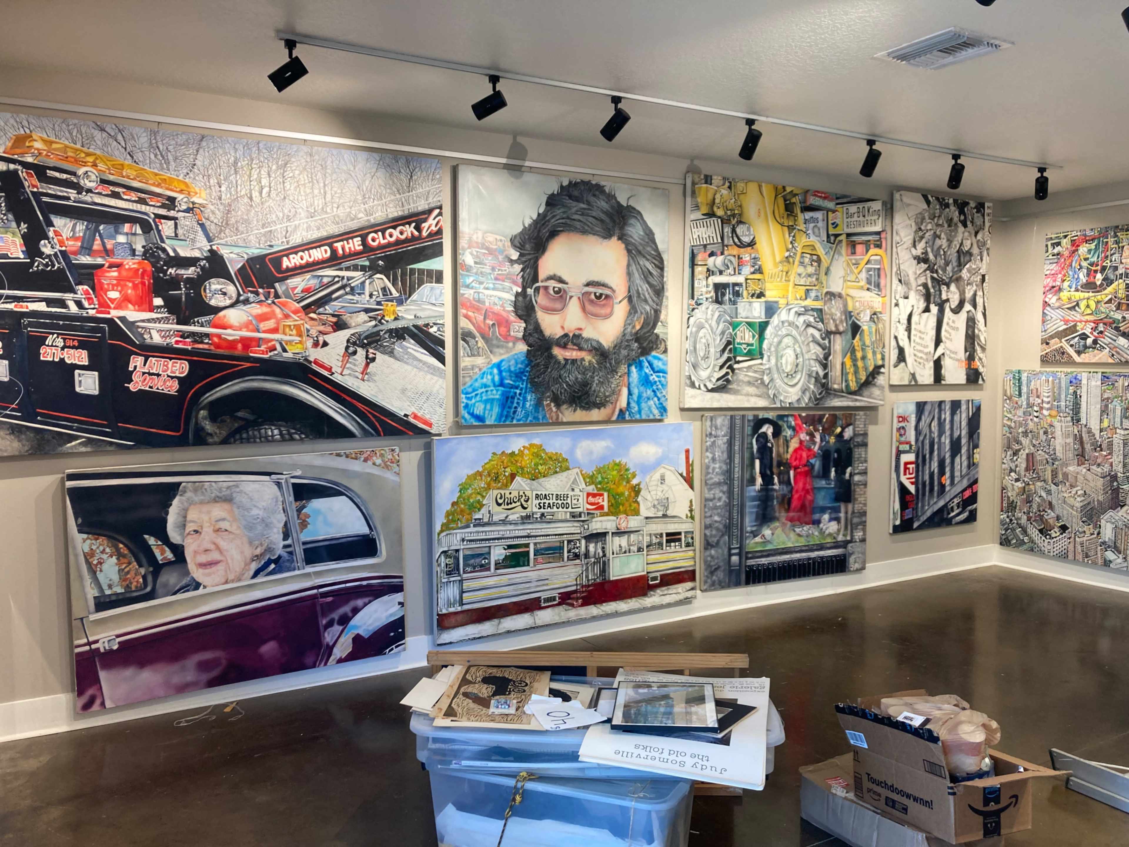 The image shows a gallery wall featuring a collection of large, colorful artworks that depict various scenes and characters, alongside boxes and containers in the foreground.