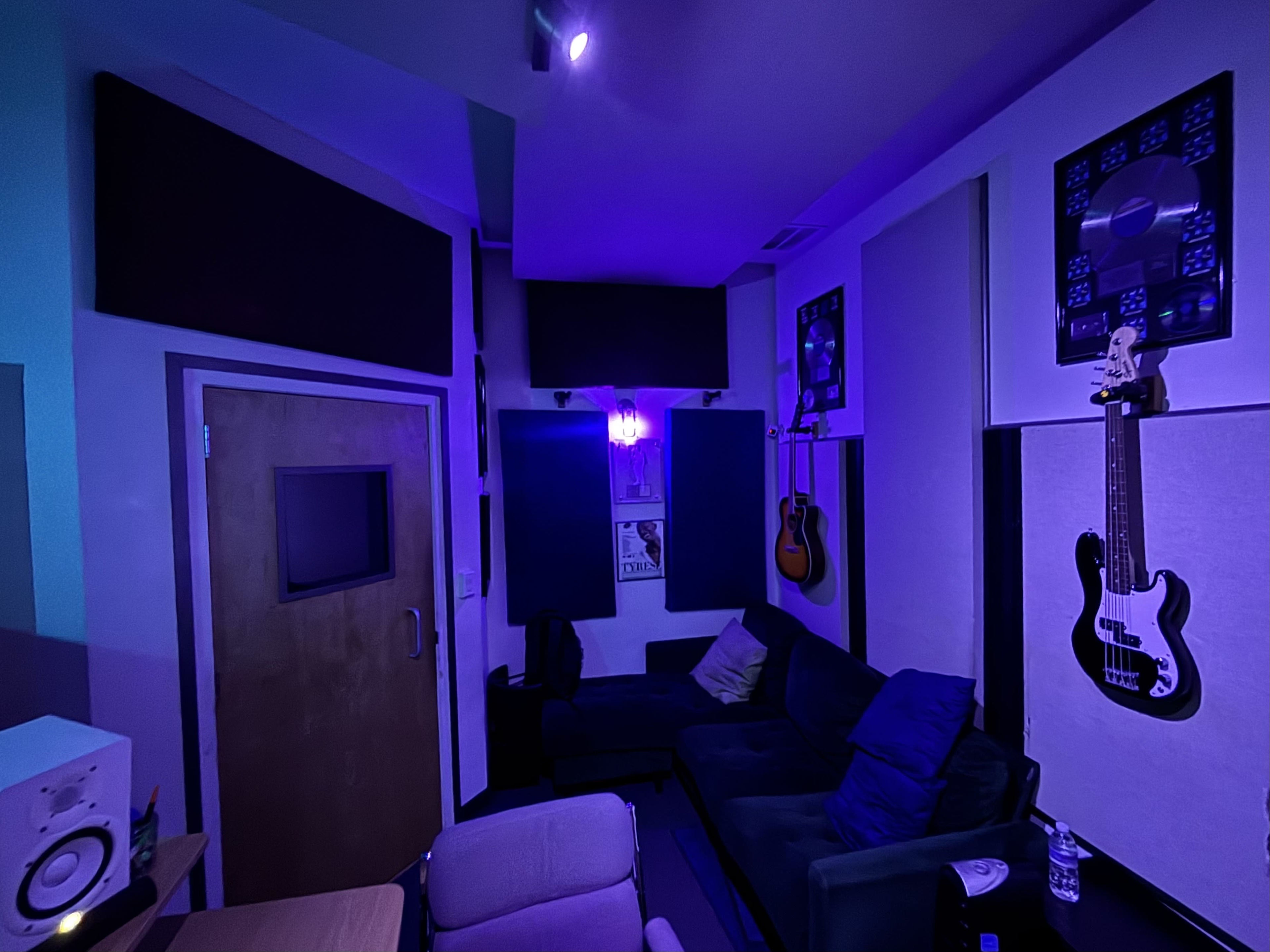 The image shows a music studio with purple lighting, featuring a seating area, guitars on the walls, soundproof panels, and an equipment setup.