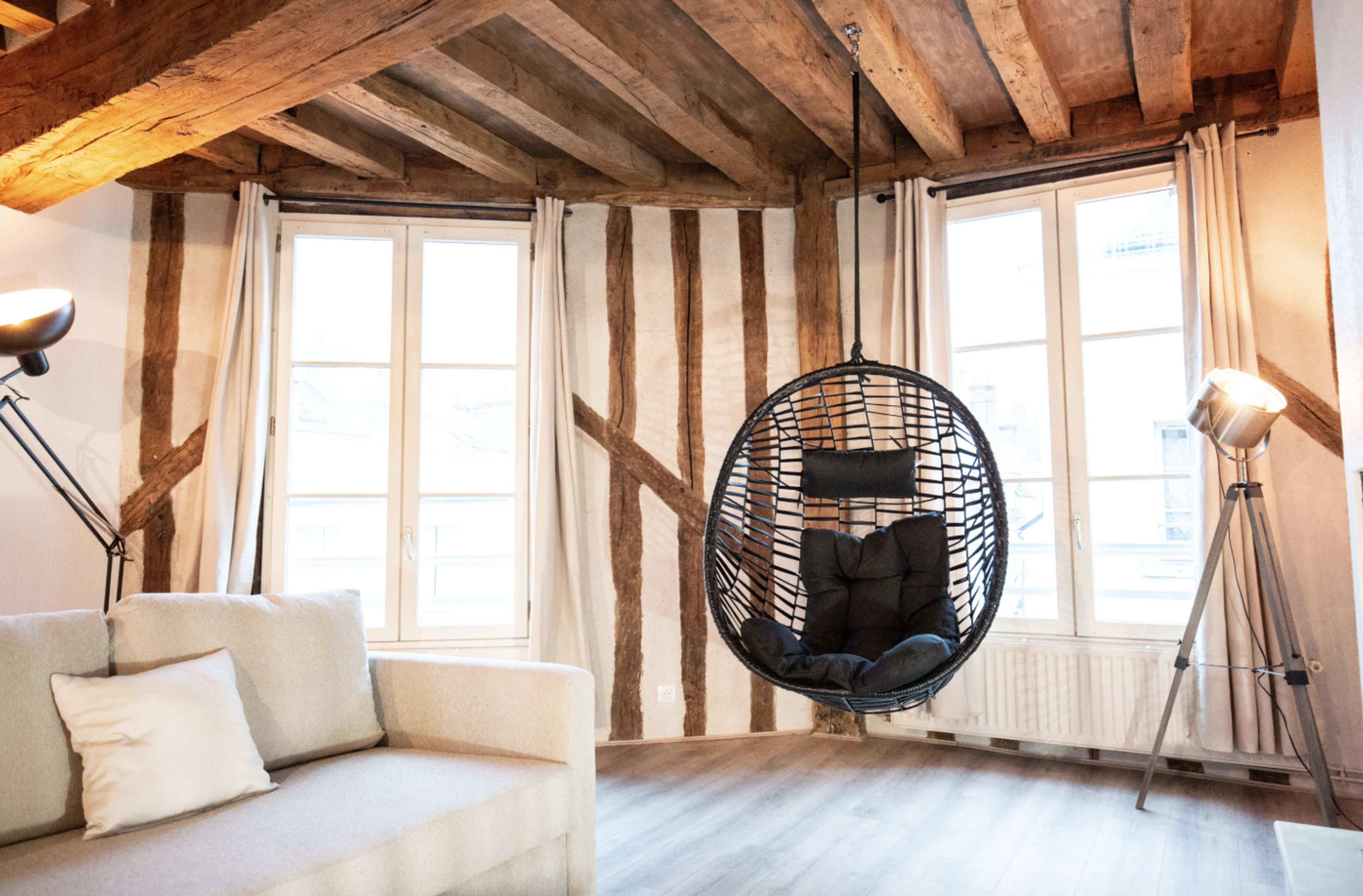 The image shows a modern living room featuring a hanging chair, a light-colored sofa, and large windows with wooden beams and striped walls.