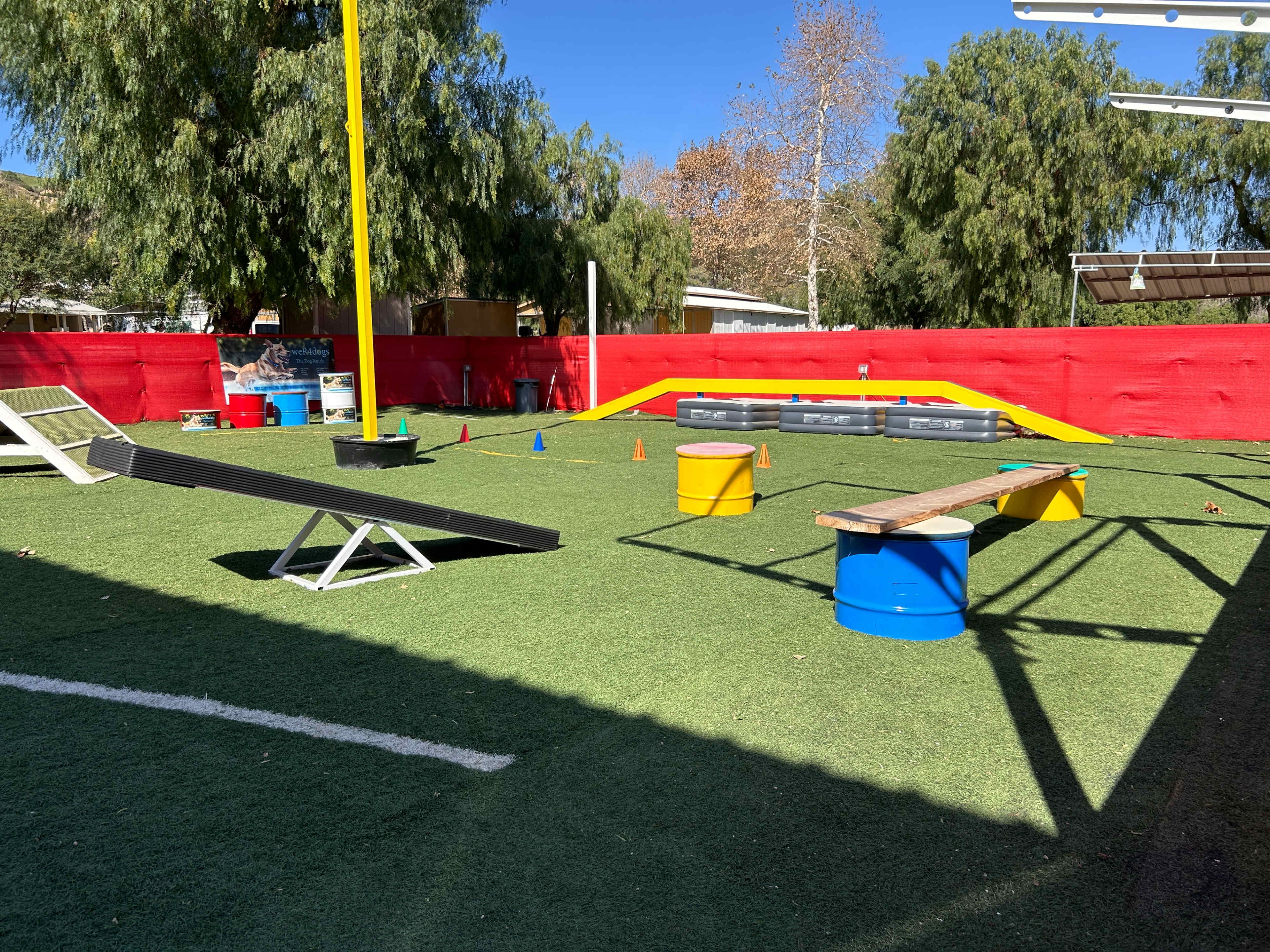 The image shows a landscaped outdoor space equipped with obstacle course elements, including ramps, balance beams, and agility cones, set on artificial turf.