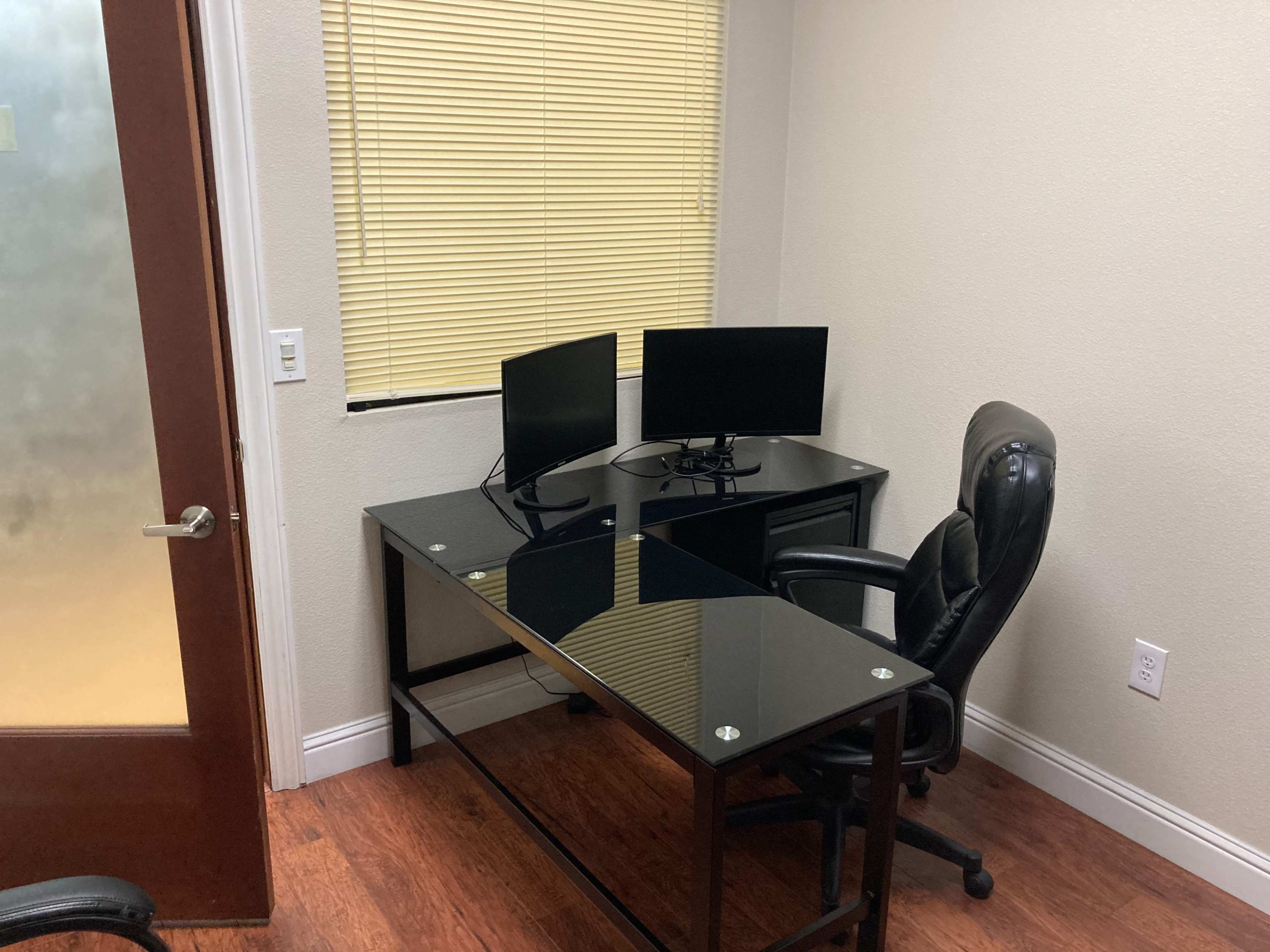 A small office with a glass-top desk, two computer monitors, a black leather office chair, and a window covered with blinds.