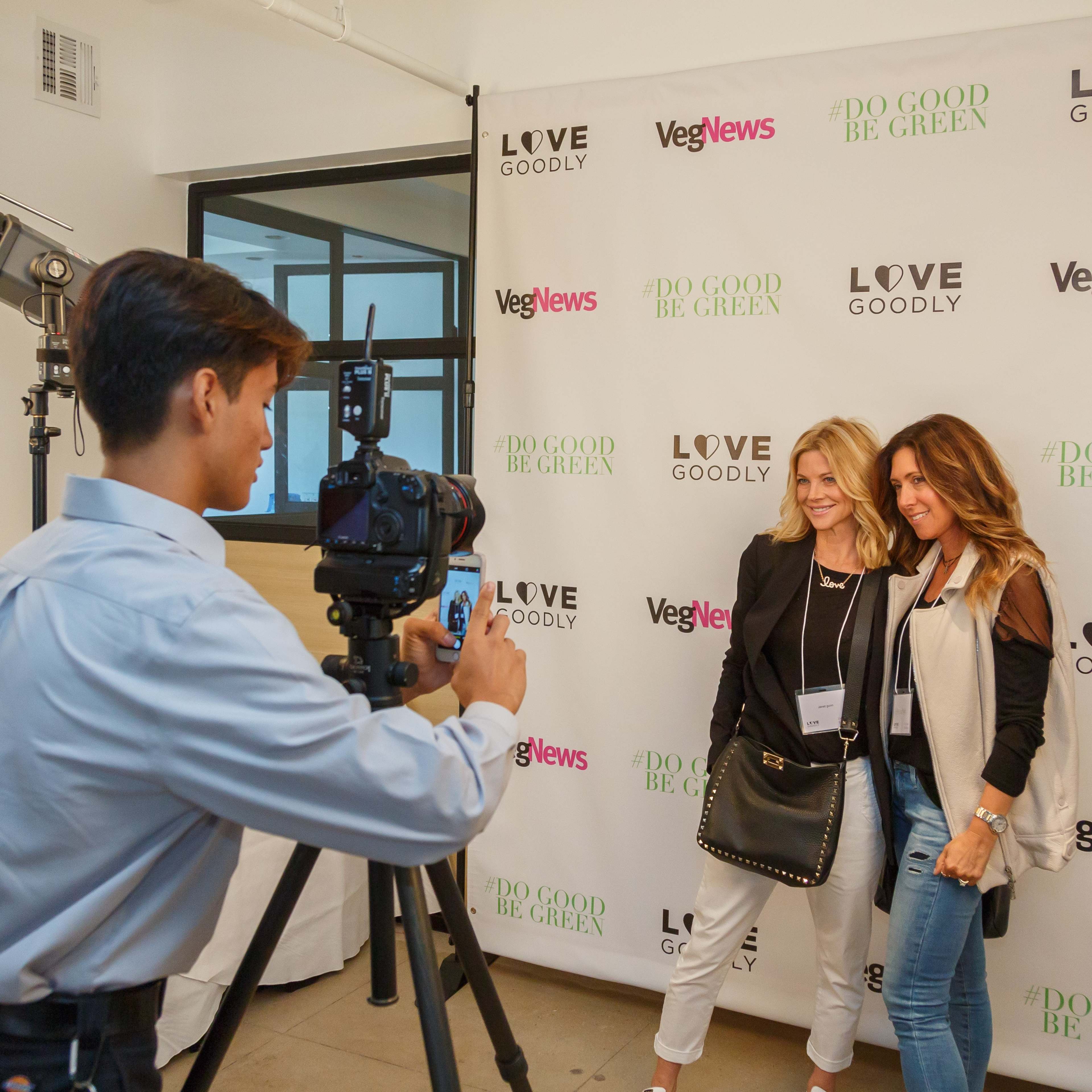 Two women pose for a photo in front of a backdrop with the logos of VegNews and Love Goodly.