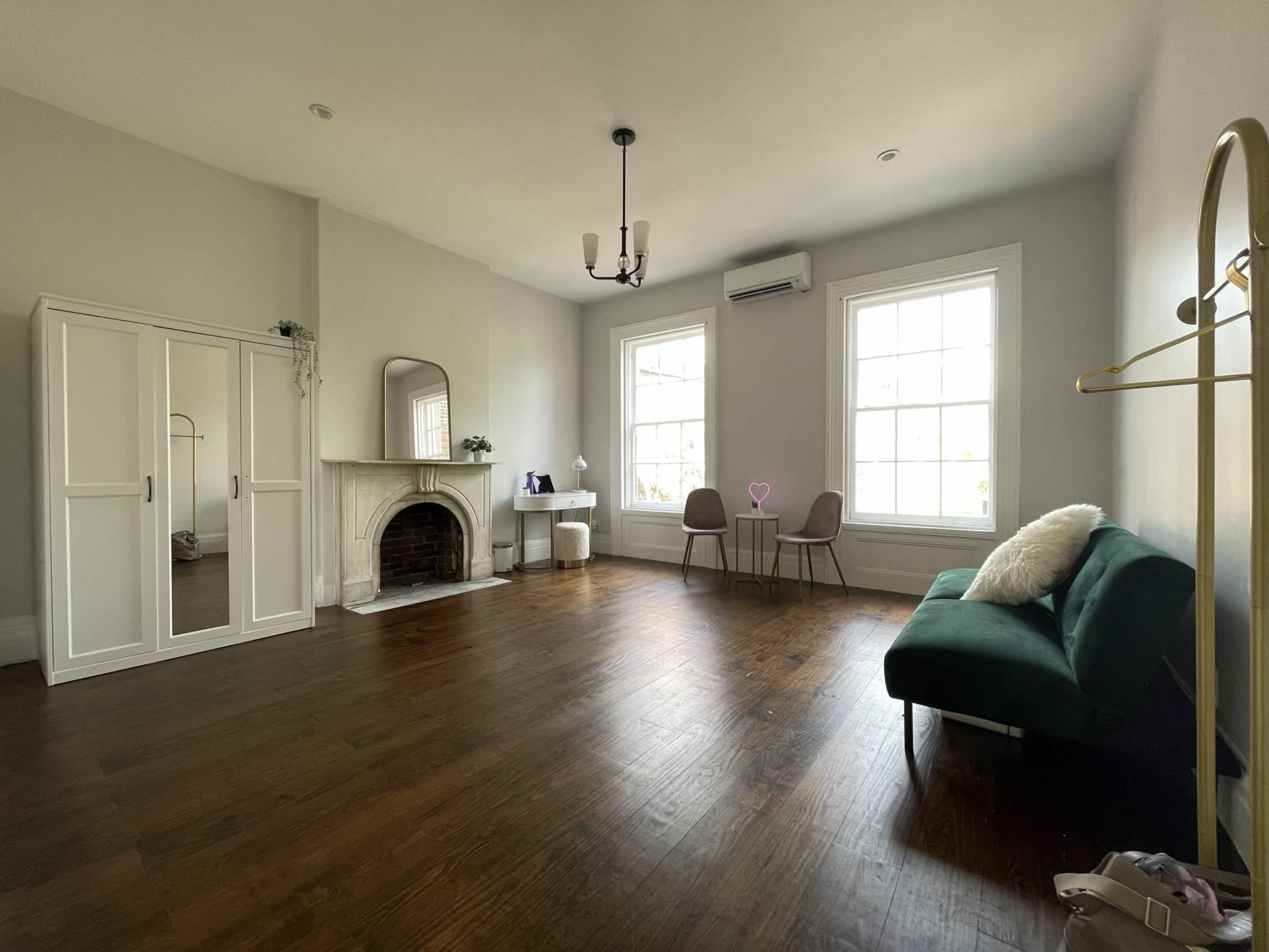 A spacious room featuring a green sofa, a dressing mirror, a fireplace, two chairs, and a small table near large windows.