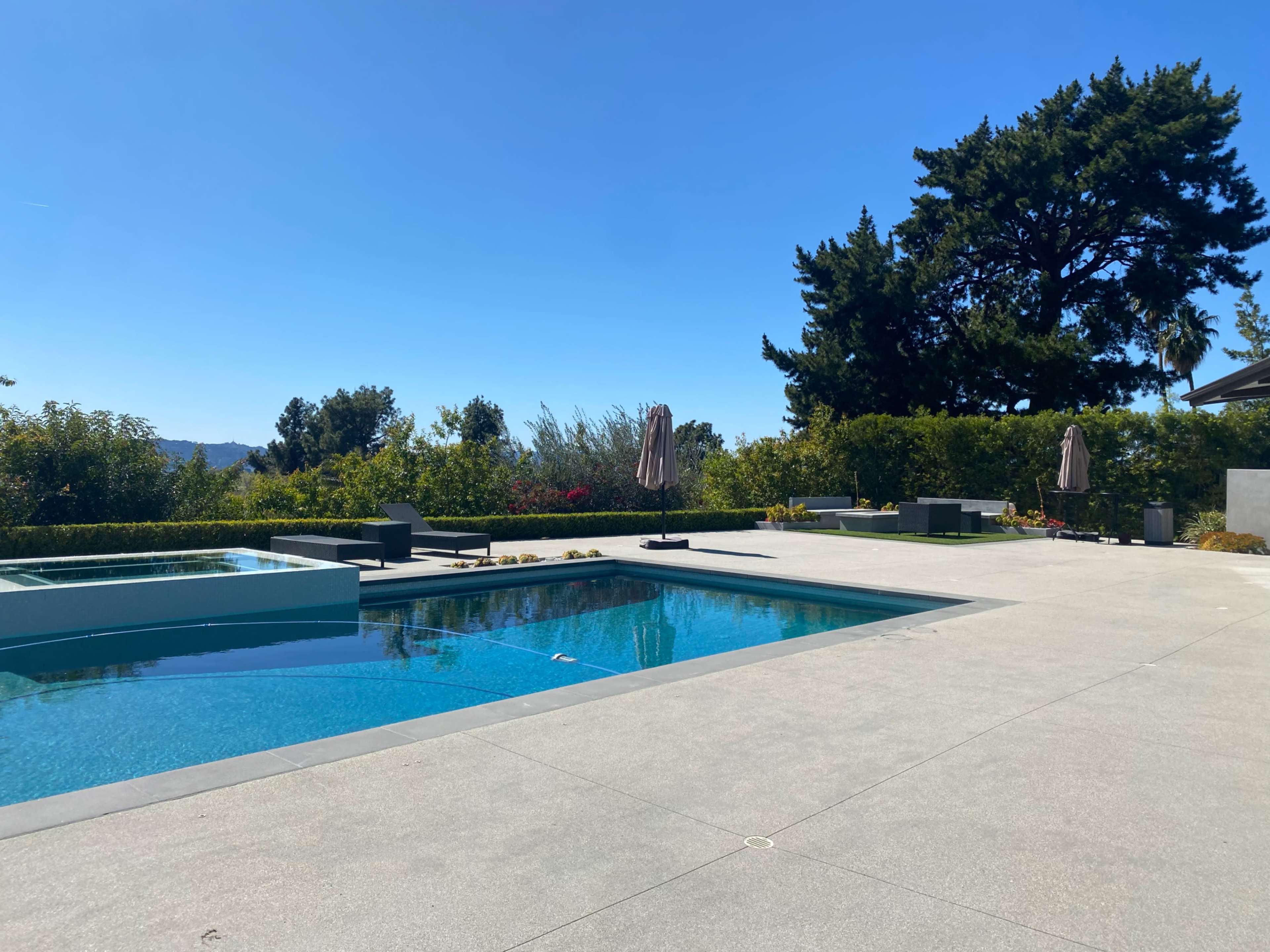 A modern outdoor pool area features a clear blue pool, lounge chairs, and landscaping under a bright blue sky.