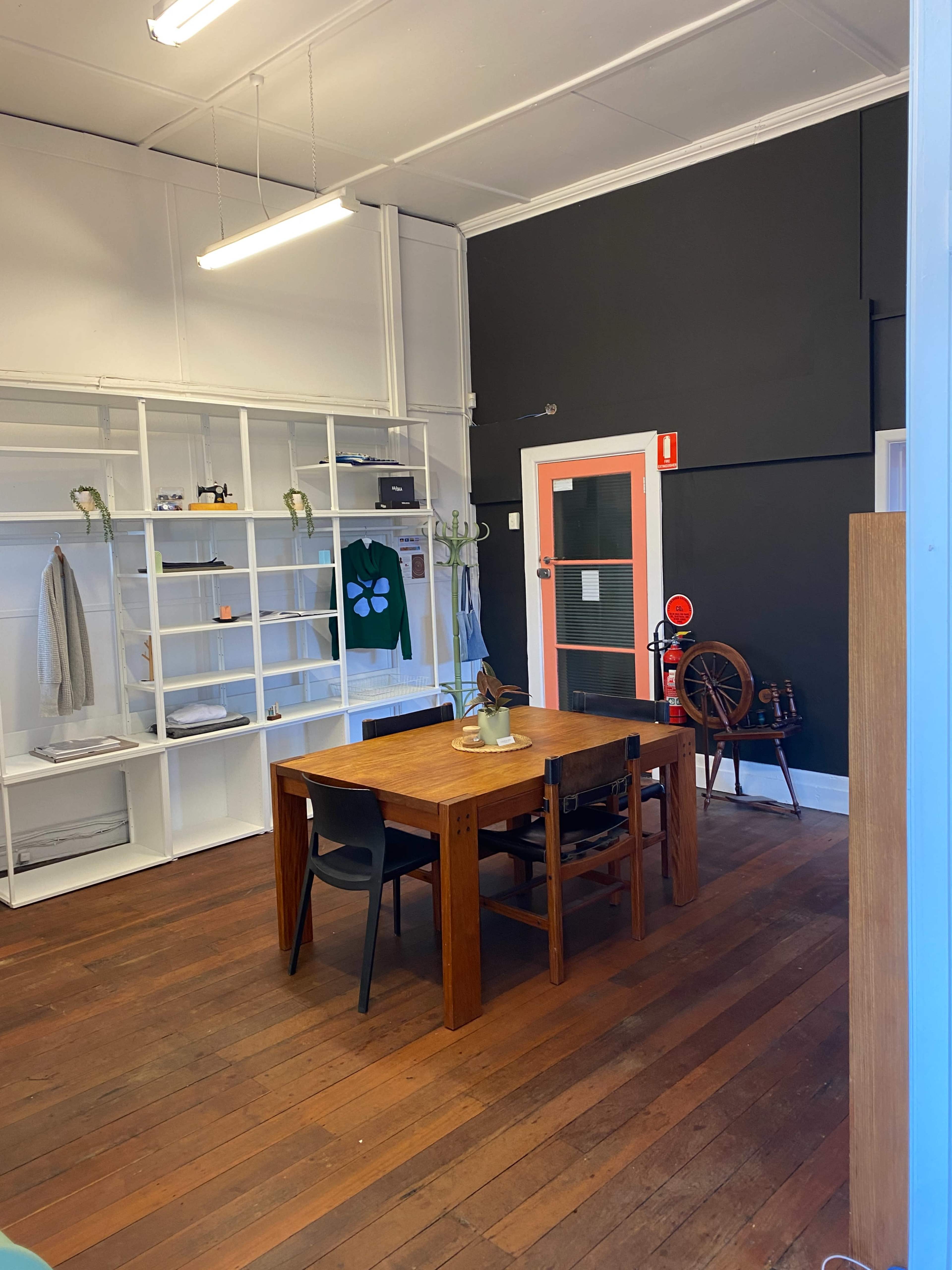 The image shows an office space with a wooden table surrounded by black chairs, shelves on the walls displaying various items, and a black wall contrasting with a bright orange door.