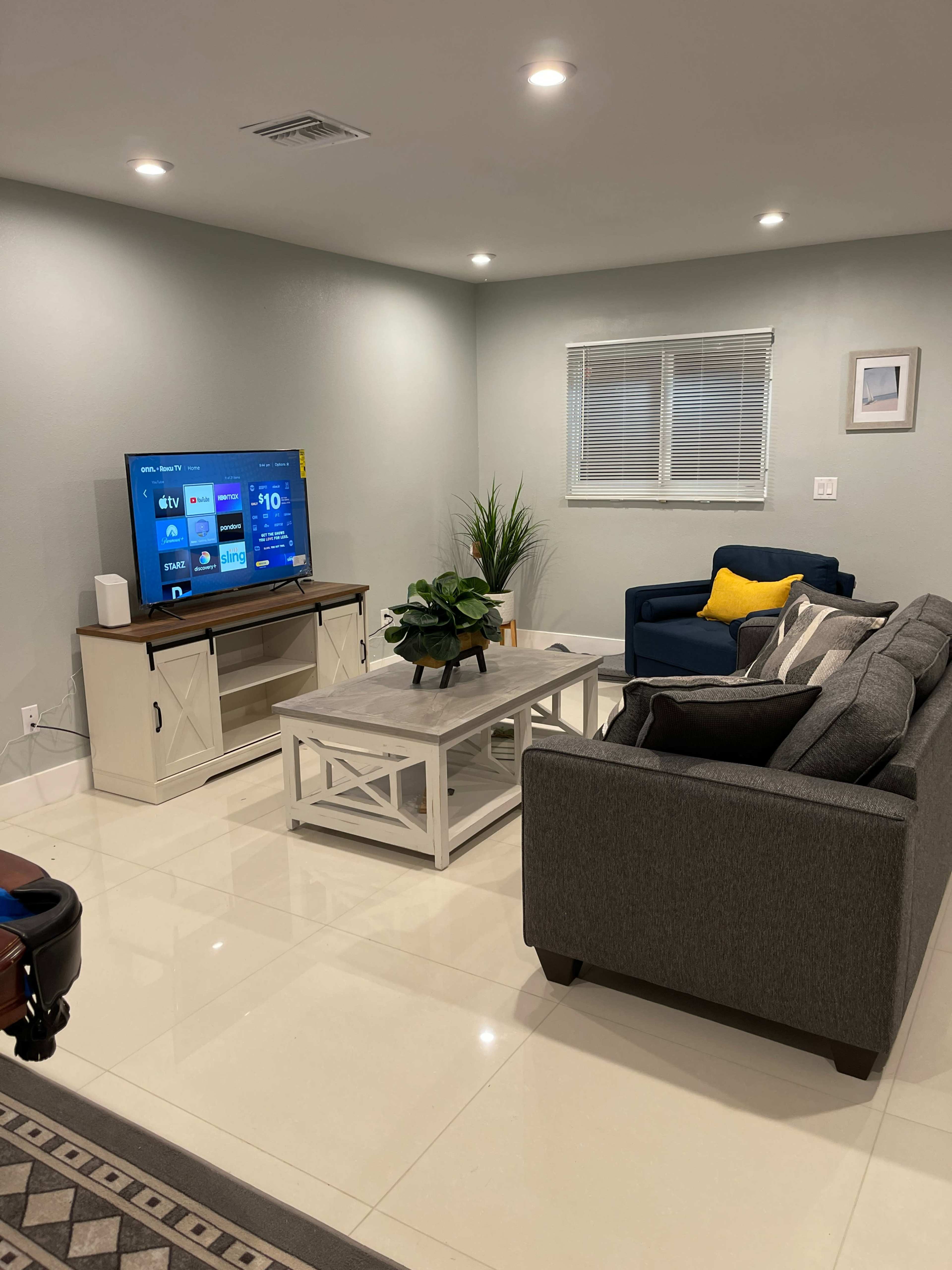 A modern living room features a gray sofa, a blue chair, a coffee table, and a television mounted on a media console with decorative plants and a window.