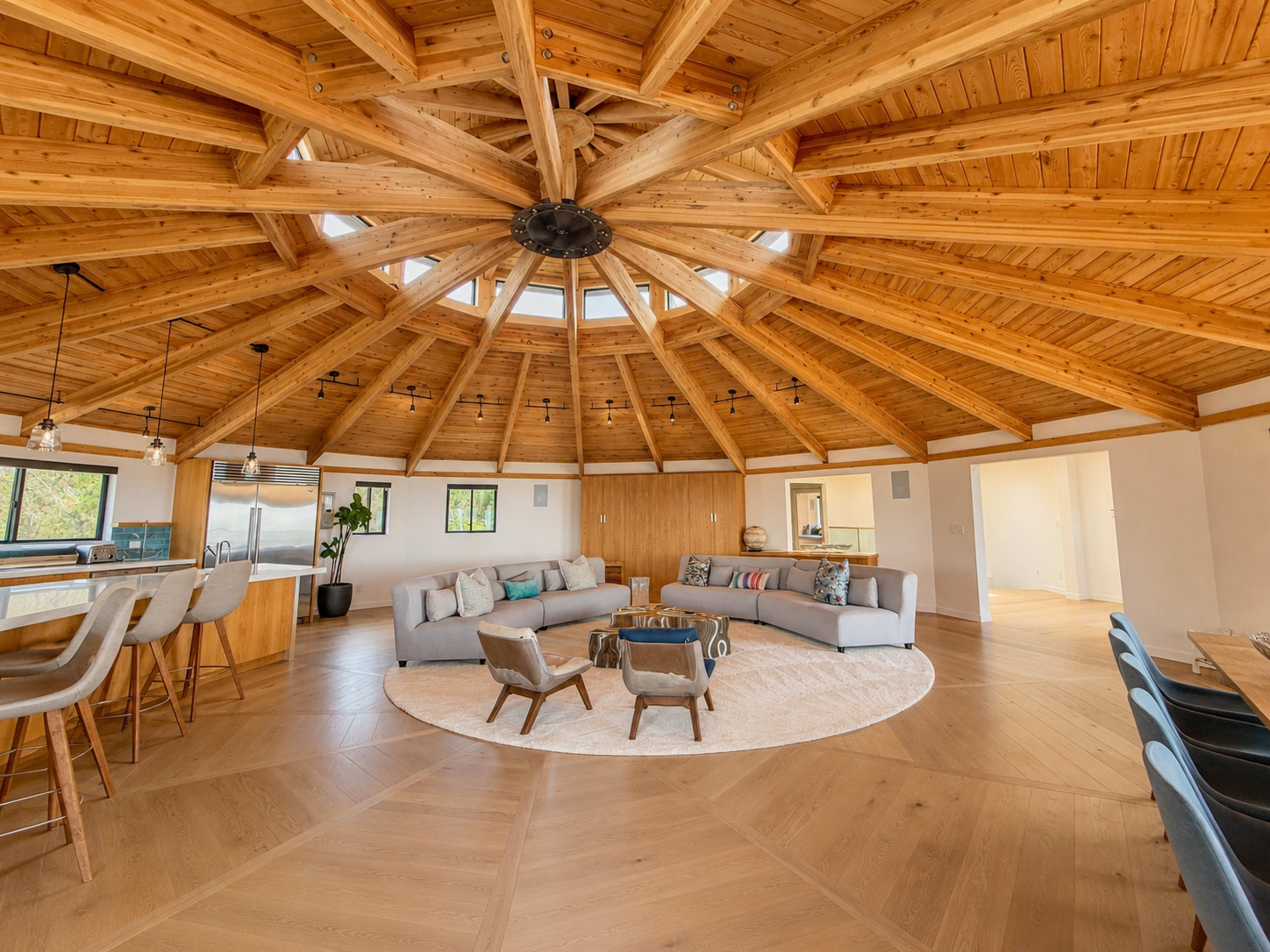 The image shows a spacious, circular living area with a vaulted wooden ceiling, a central seating arrangement on a circular rug, and large windows allowing natural light to enter.