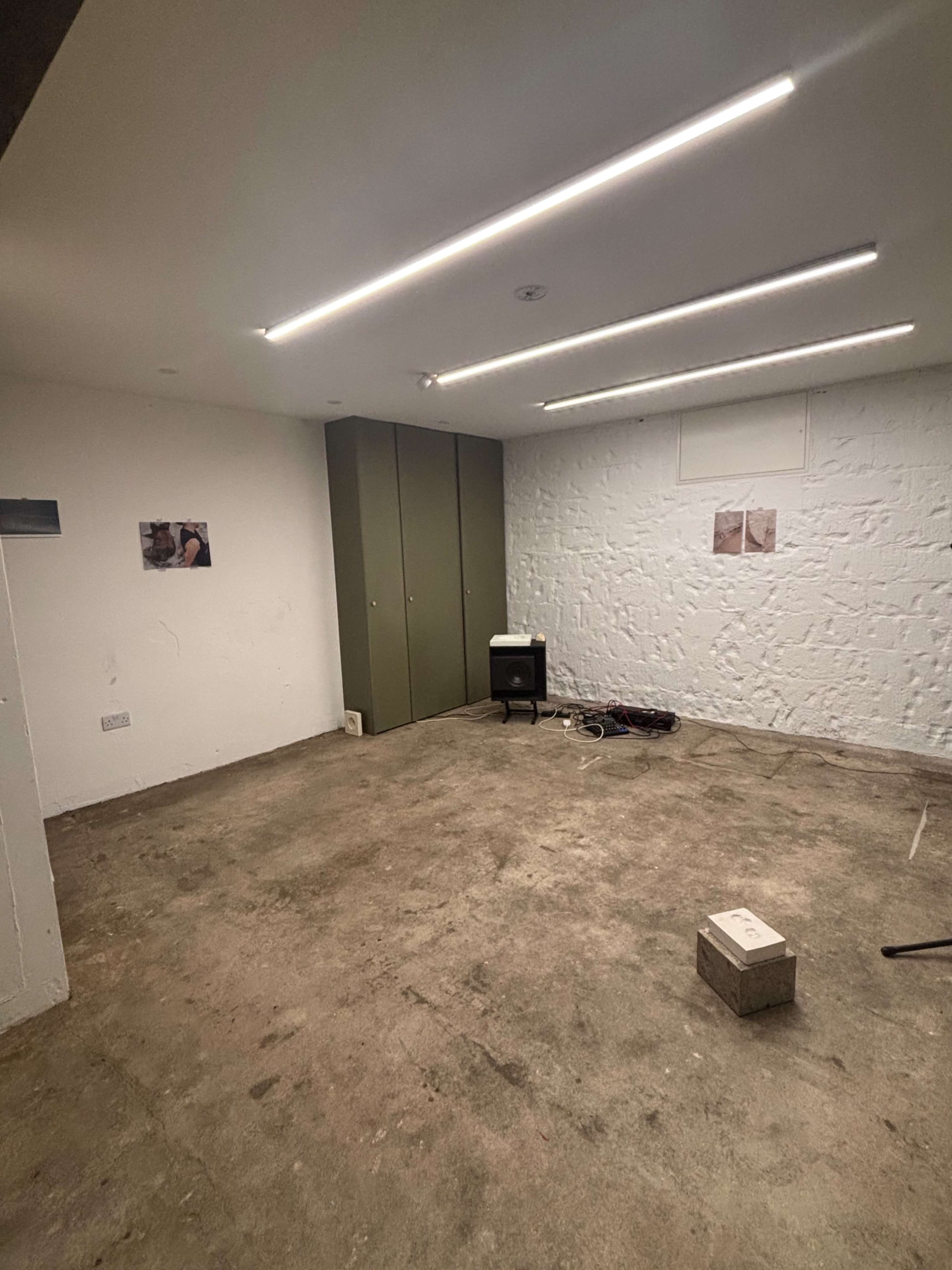 The image shows an empty, unfinished room with a concrete floor, white walls, a cabinet against one wall, and two photographs hanging on the opposite wall.