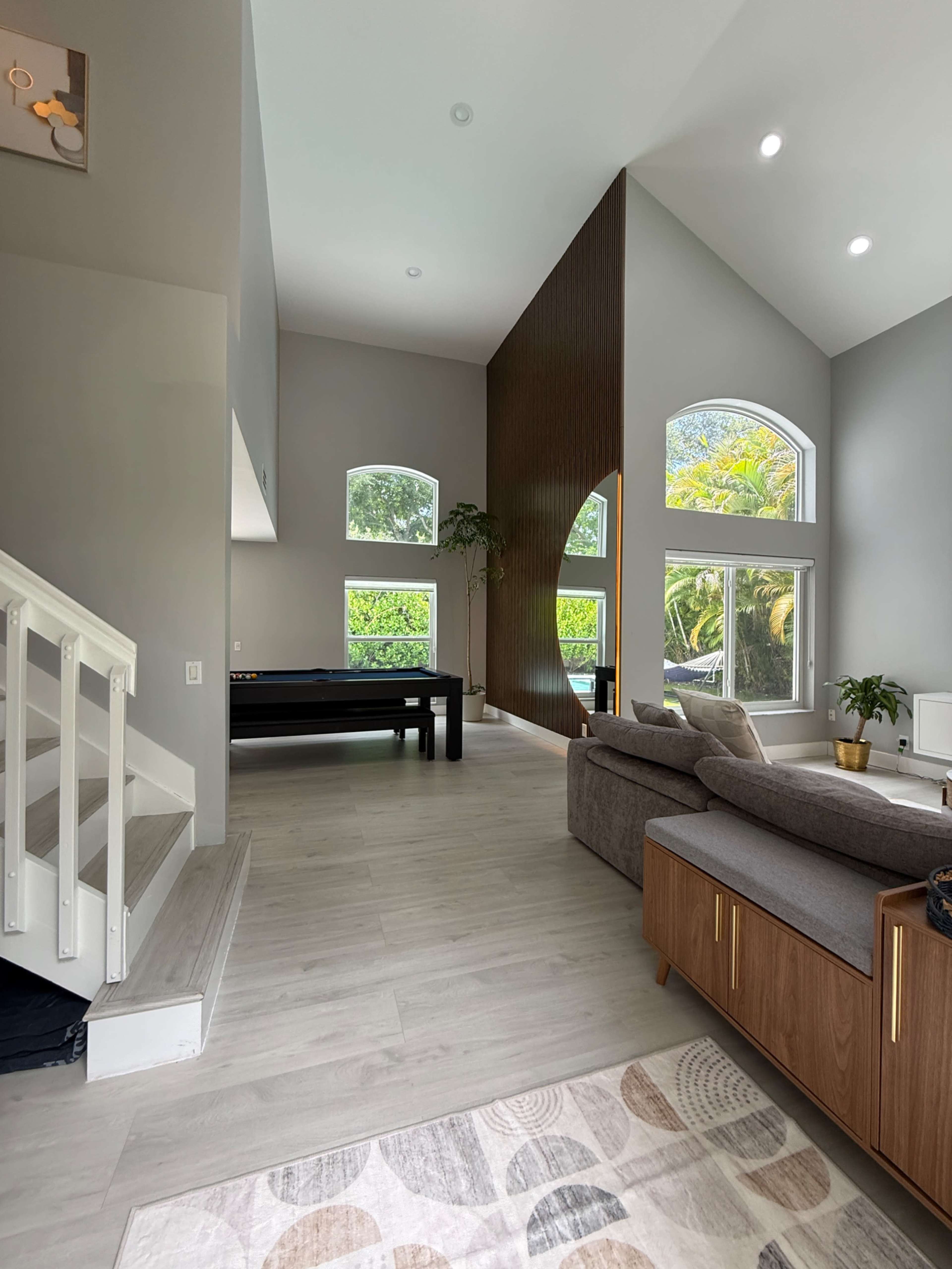 The image shows a spacious living area with high ceilings, large windows, a comfortable sectional sofa, and a pool table in the corner.