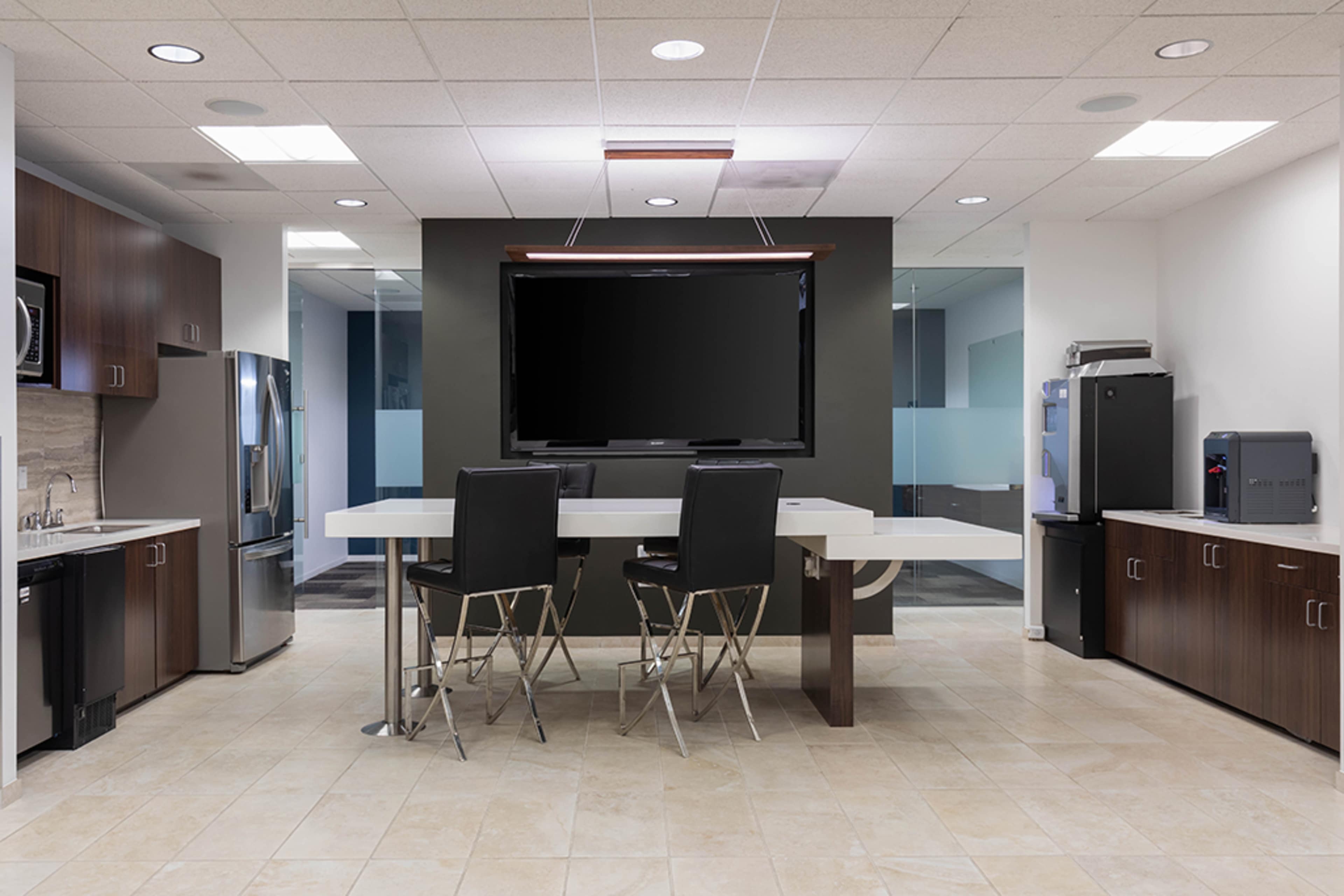 A modern office kitchenette with a large table, black chairs, and a flat-screen TV mounted on the wall.