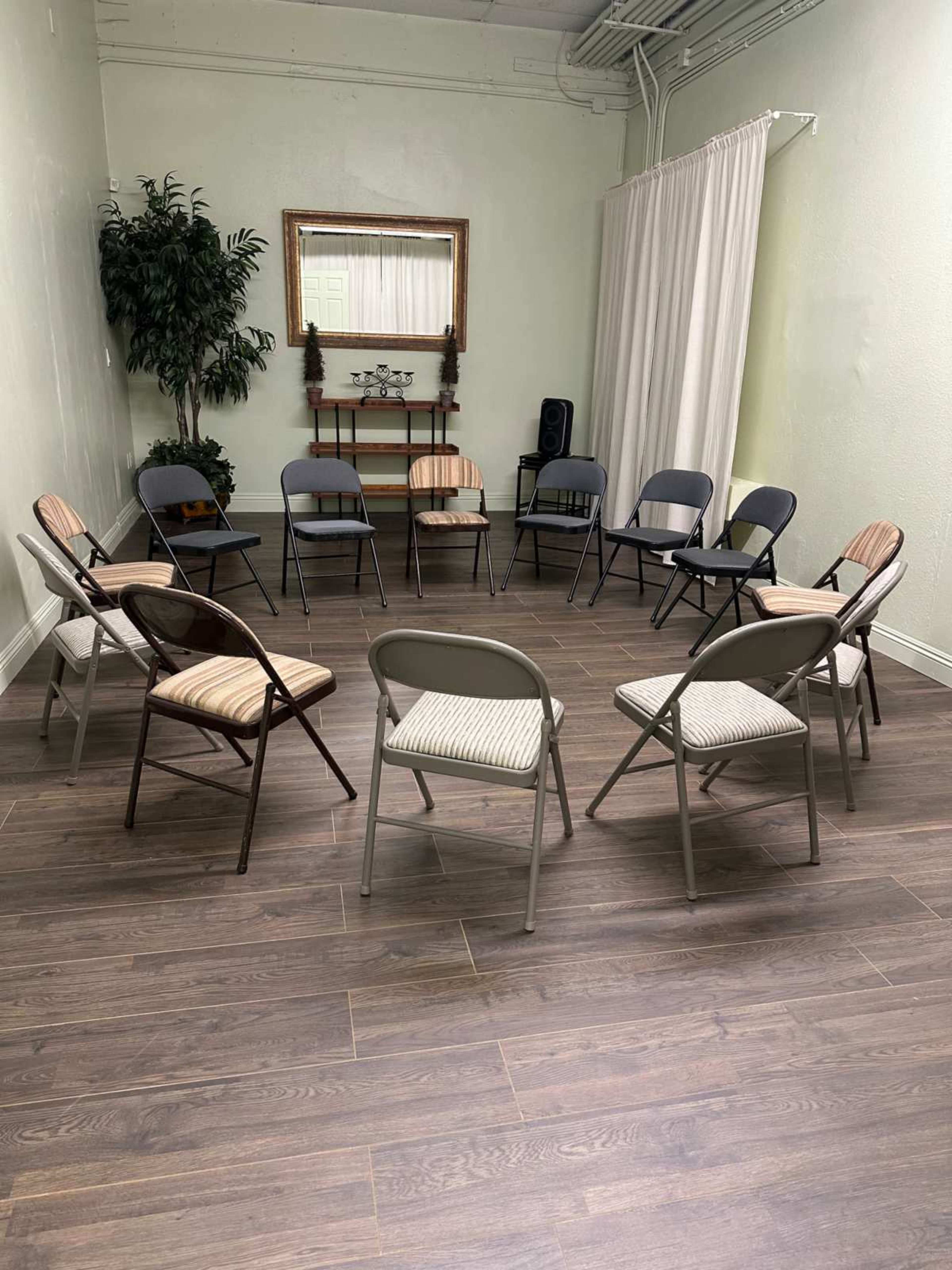 A circular arrangement of folding chairs is set up in a well-lit room with a plant and a mirror in the background.