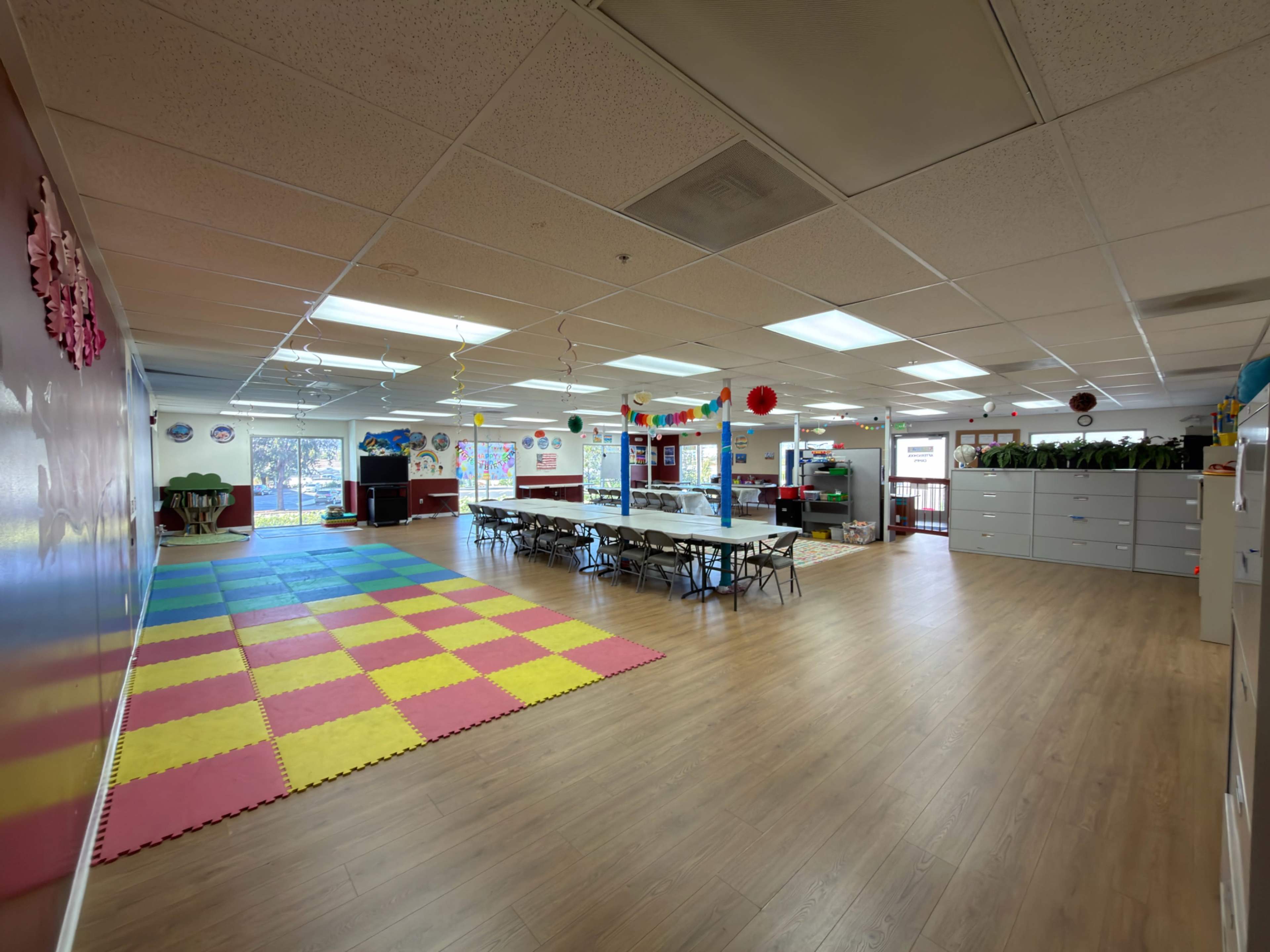 The image shows a spacious classroom with colorful rubber tiles on the floor, arranged tables for activities, and various educational decorations on the walls.