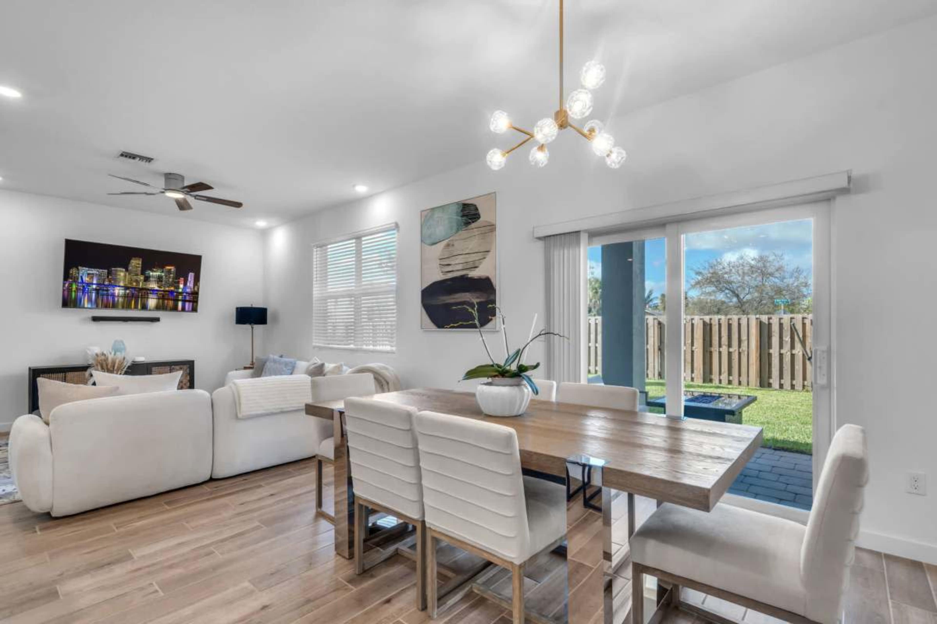 The image shows a modern dining area with a wooden table surrounded by white chairs, a sectional sofa, a television mounted on the wall, and sliding glass doors leading to an outdoor space.