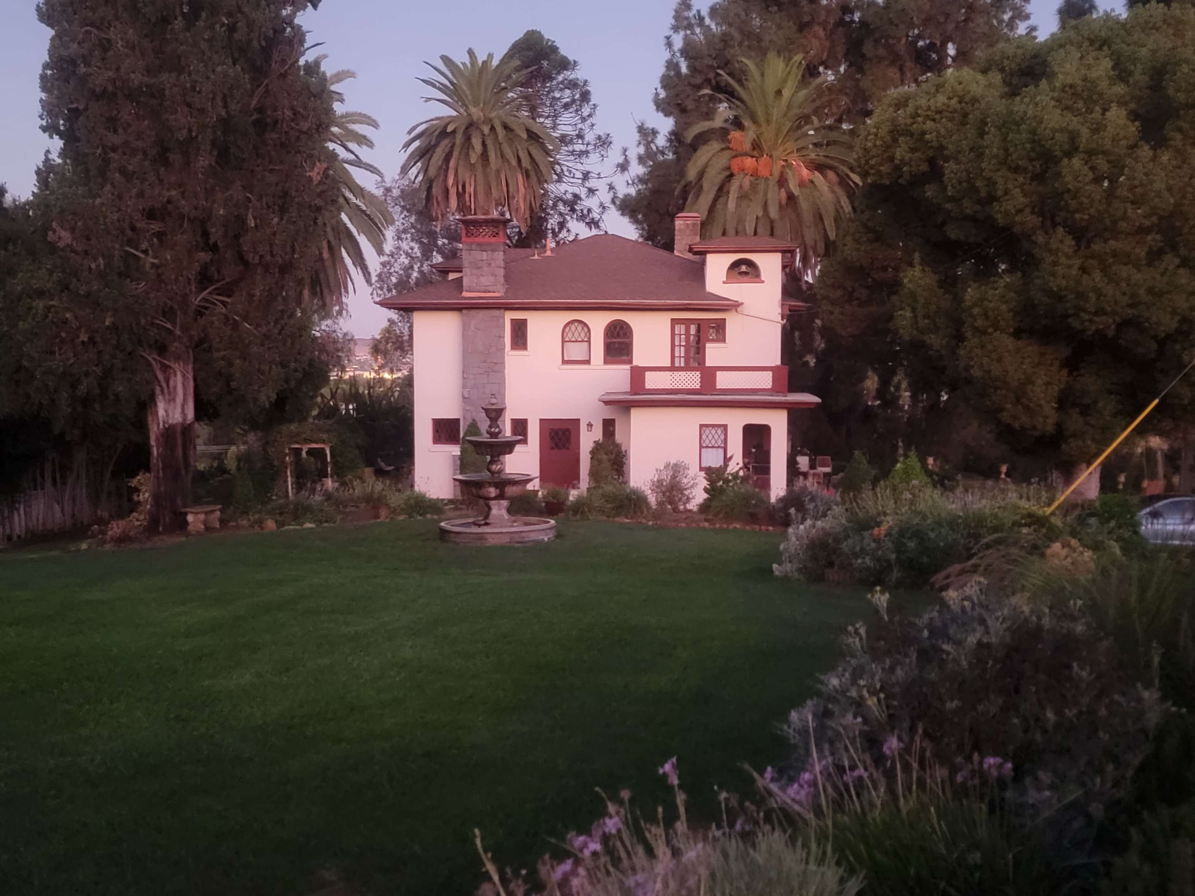 A two-story white house with a red roof and palm trees stands in a lush green yard, featuring a stone fountain and various plants.