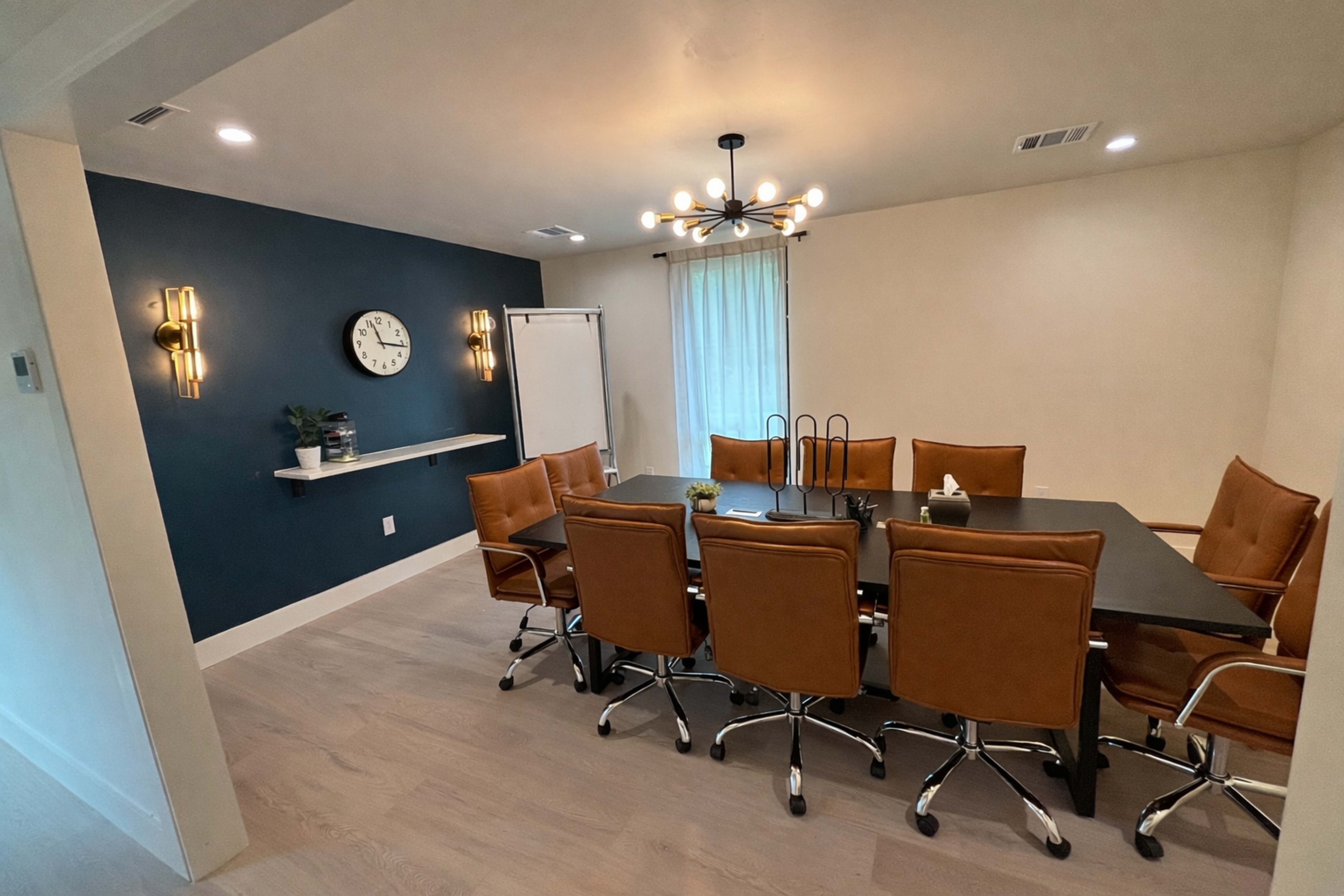 The image shows a modern conference room with a long black table surrounded by brown leather chairs, a wall clock, and a whiteboard on display.