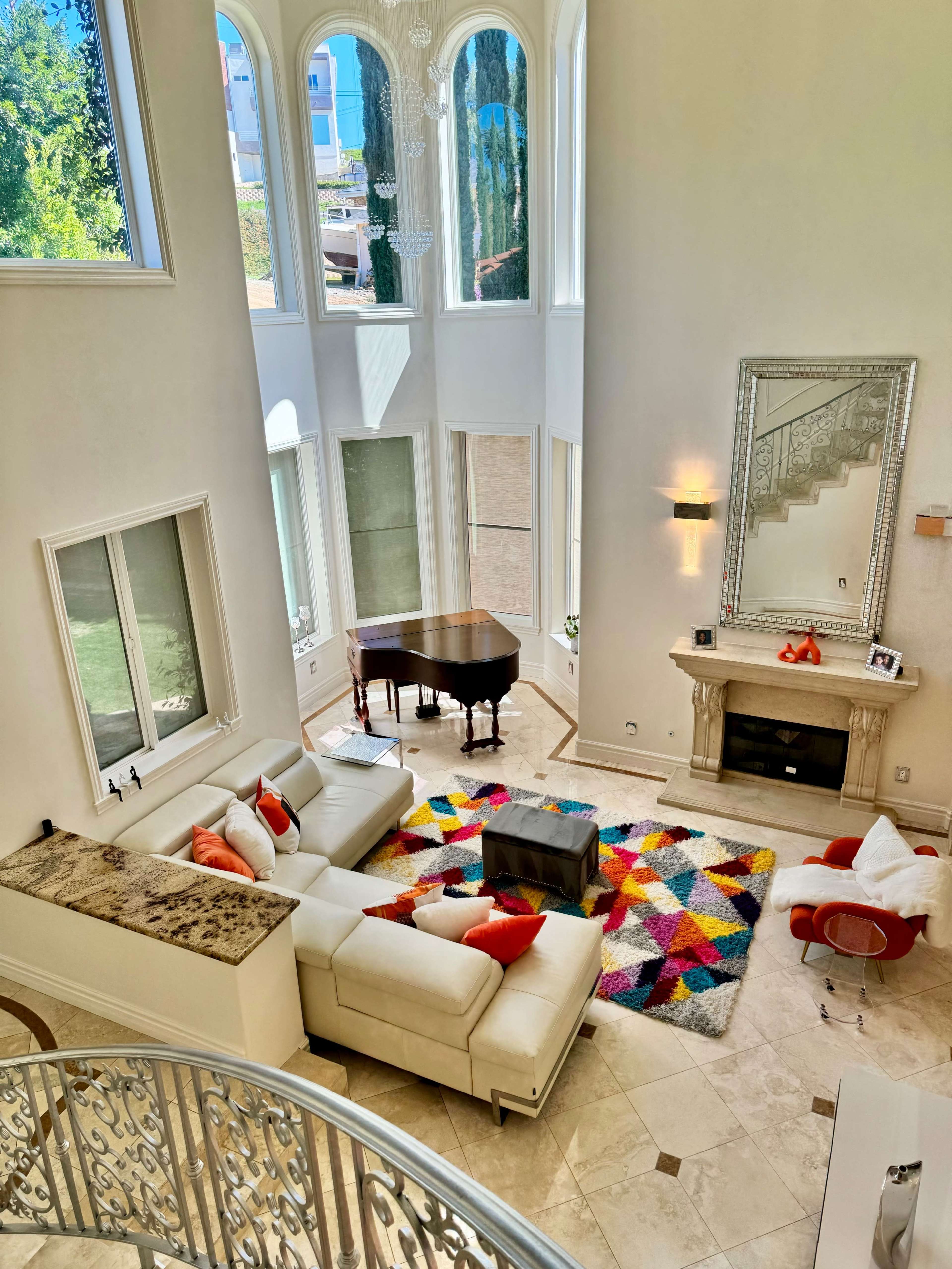 A spacious living room features large windows, a grand piano, a white sectional sofa, and a colorful area rug on marble flooring.