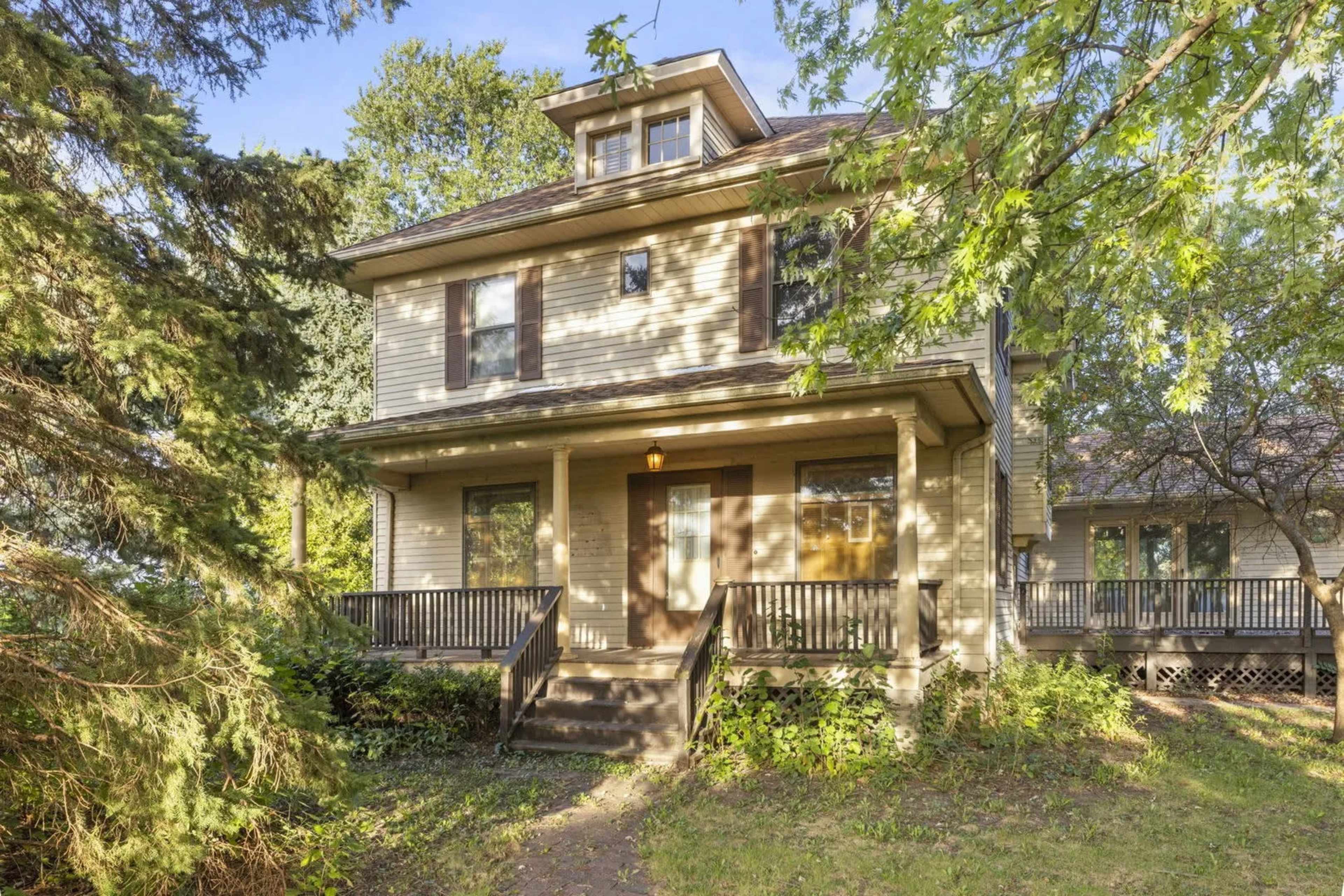 A two-story house with a sloped roof and front porch is surrounded by trees and greenery.
