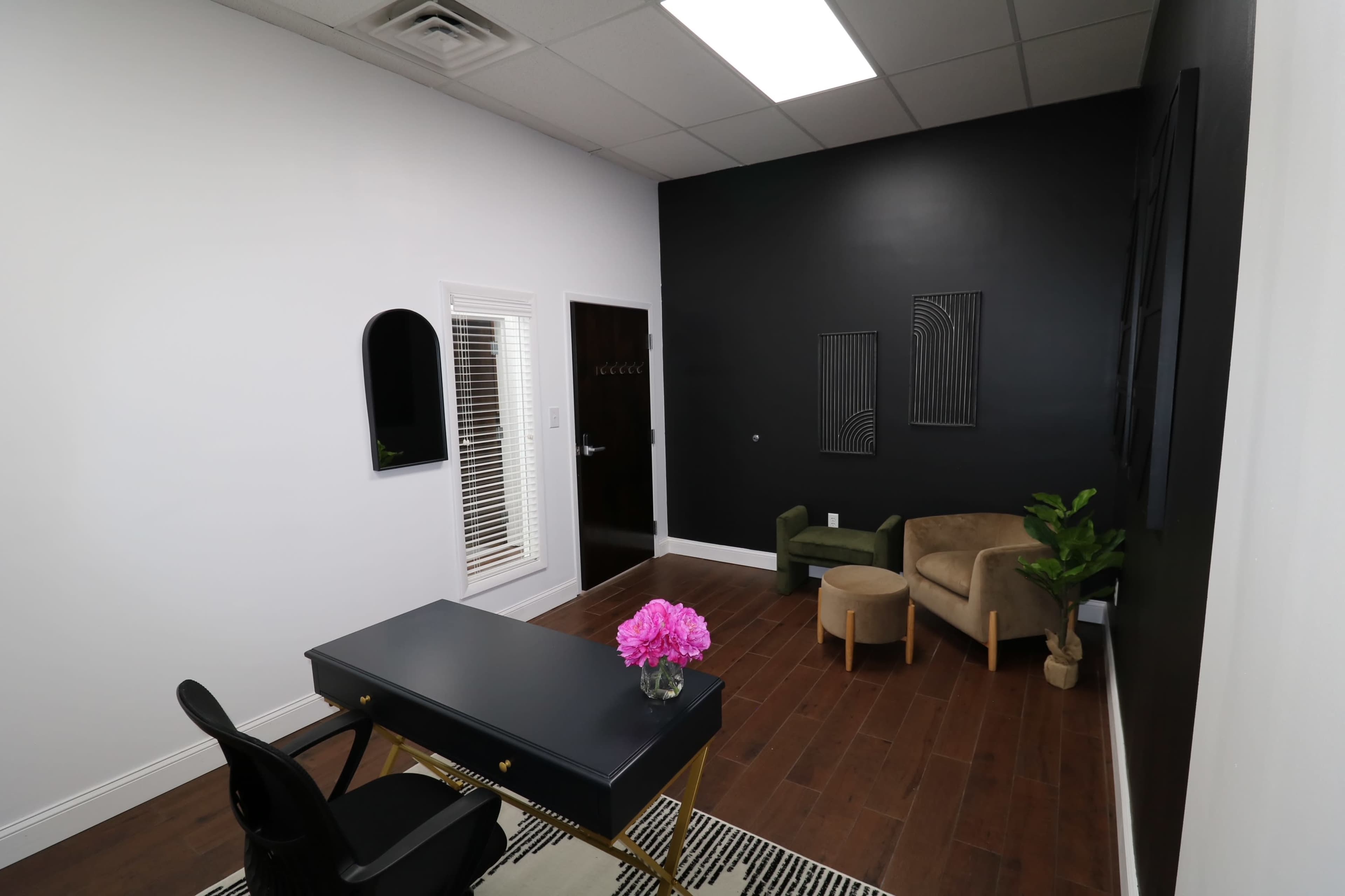 A modern office space with a black accent wall, a wooden floor, a black desk with a flower vase, and a small seating area with two chairs.