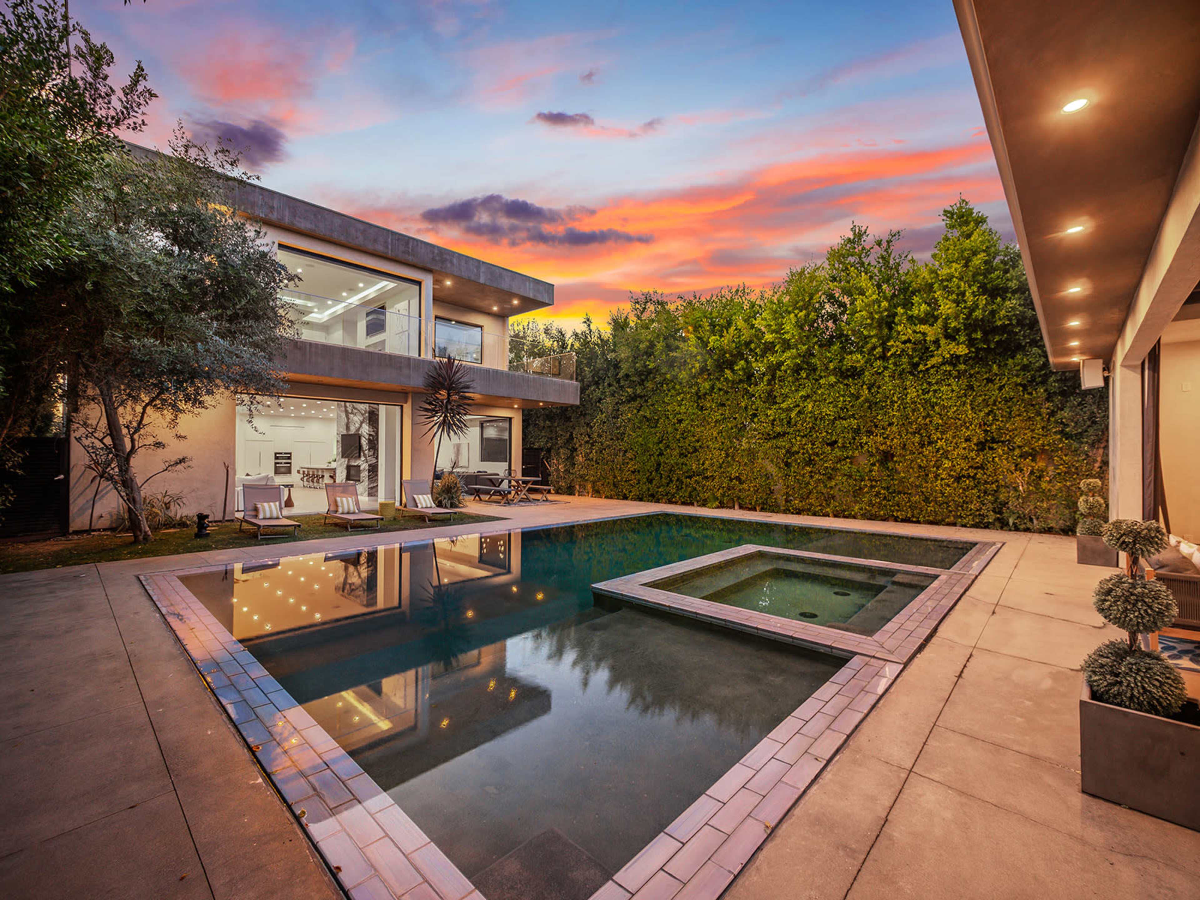 A modern house with large windows, a swimming pool, and a spa area is surrounded by greenery at sunset.