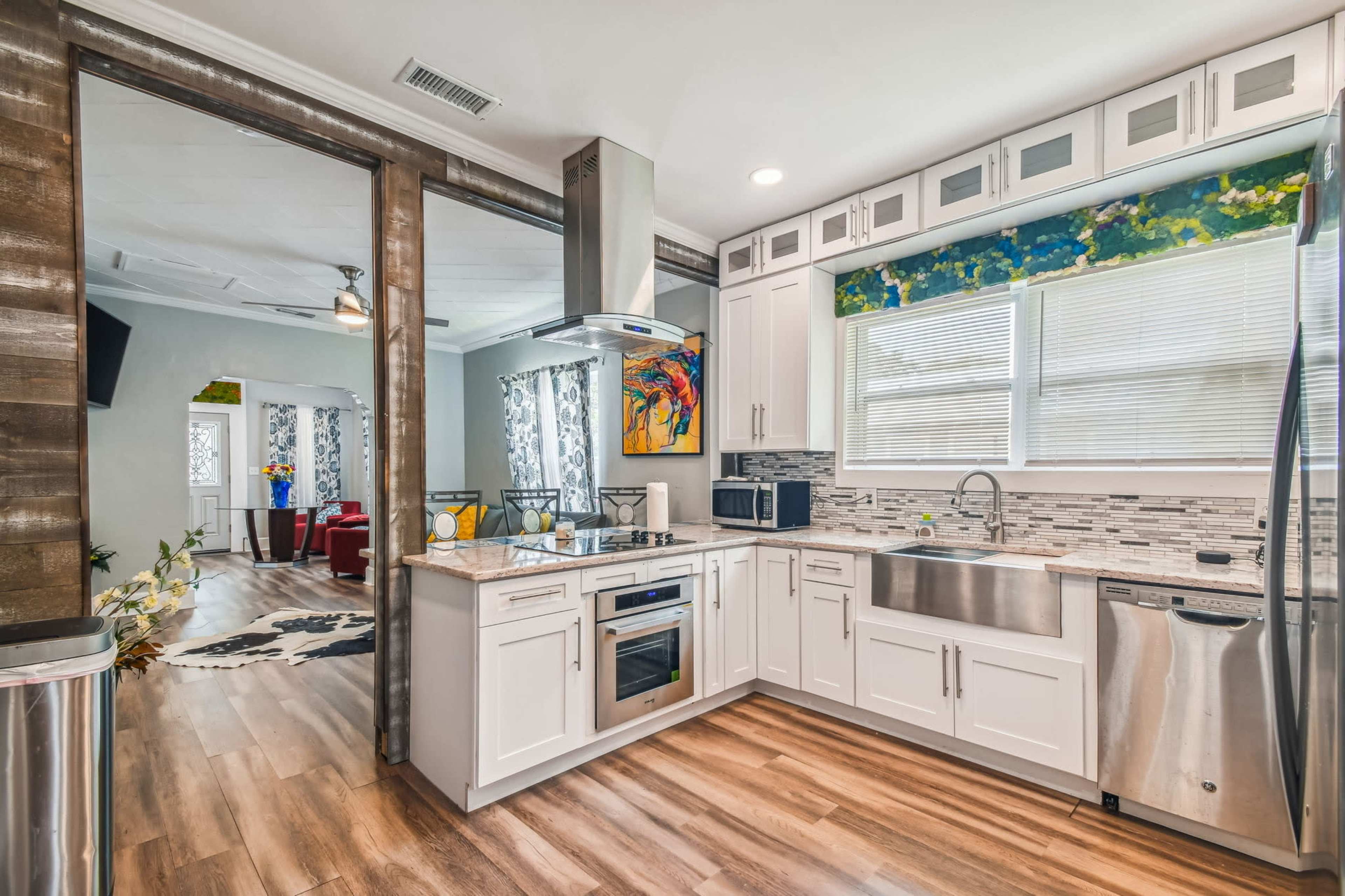 A modern kitchen features white cabinetry, stainless steel appliances, and a countertop with a large sink, adjacent to a living area with colorful decor.
