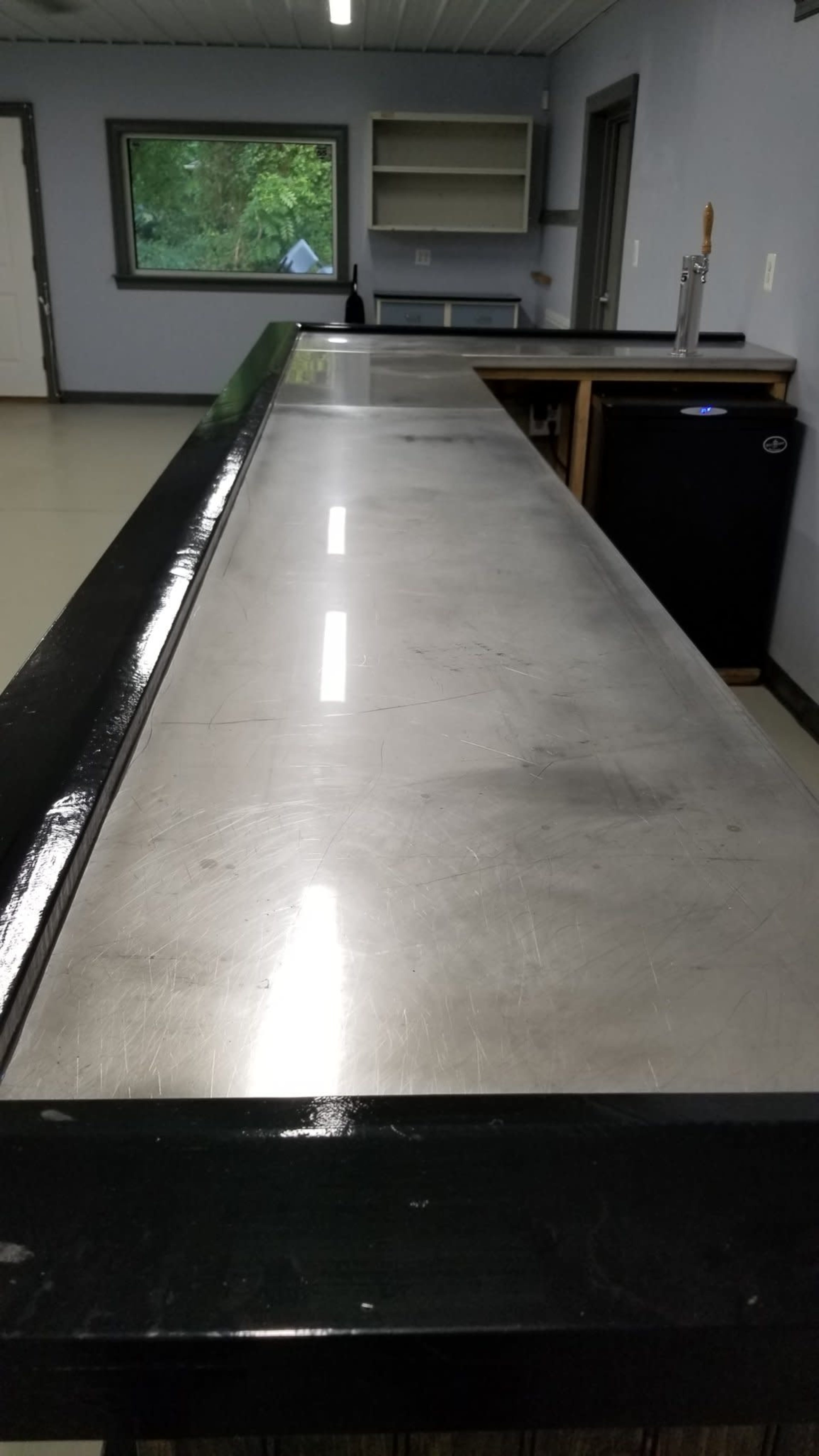 A long, stainless steel countertop in an empty room with a window and a small refrigerator in the corner.