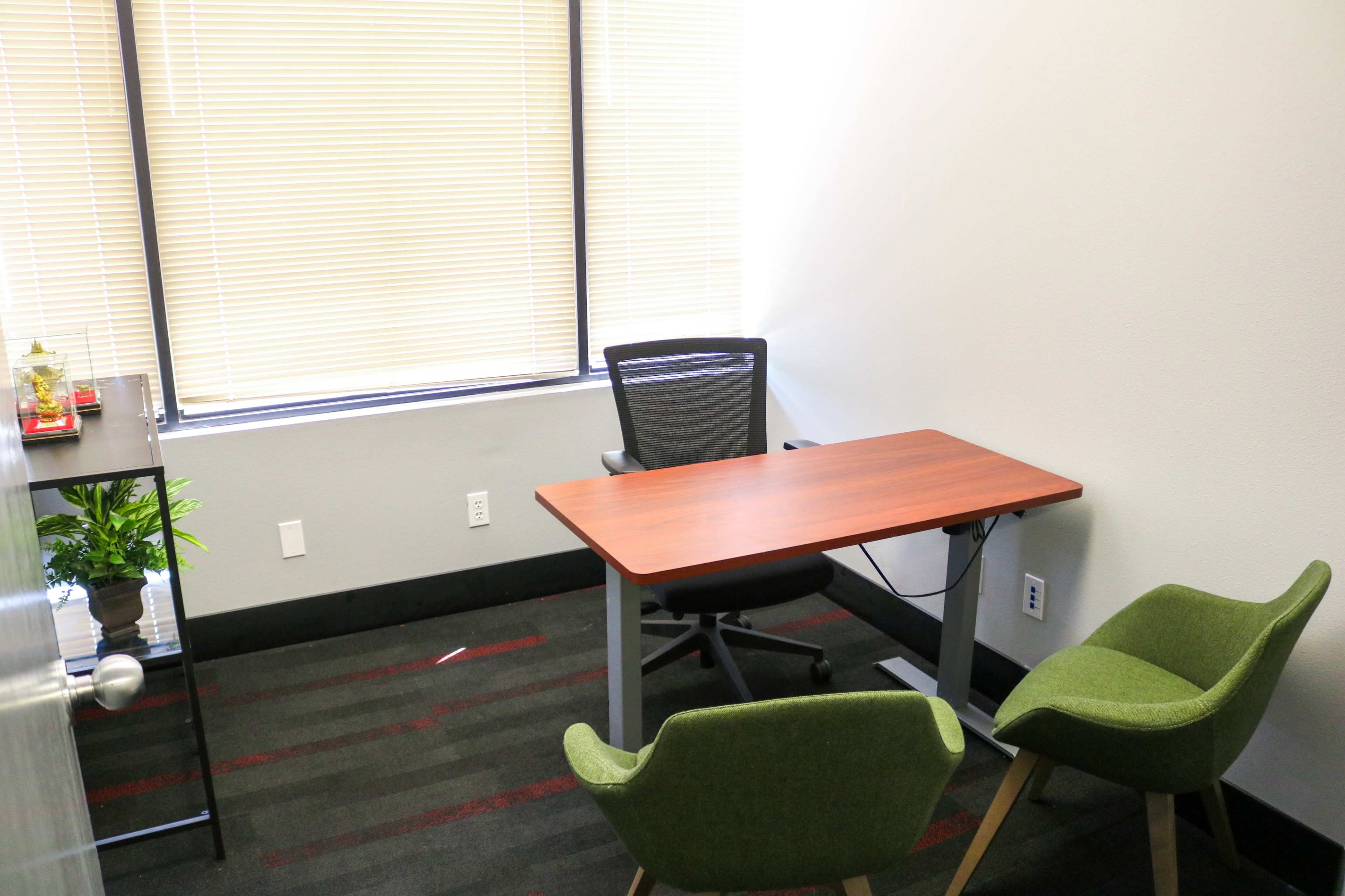 The image shows a small office space featuring a wooden desk, a rolling chair, and two green armchairs, with a potted plant and vertical blinds letting in natural light.