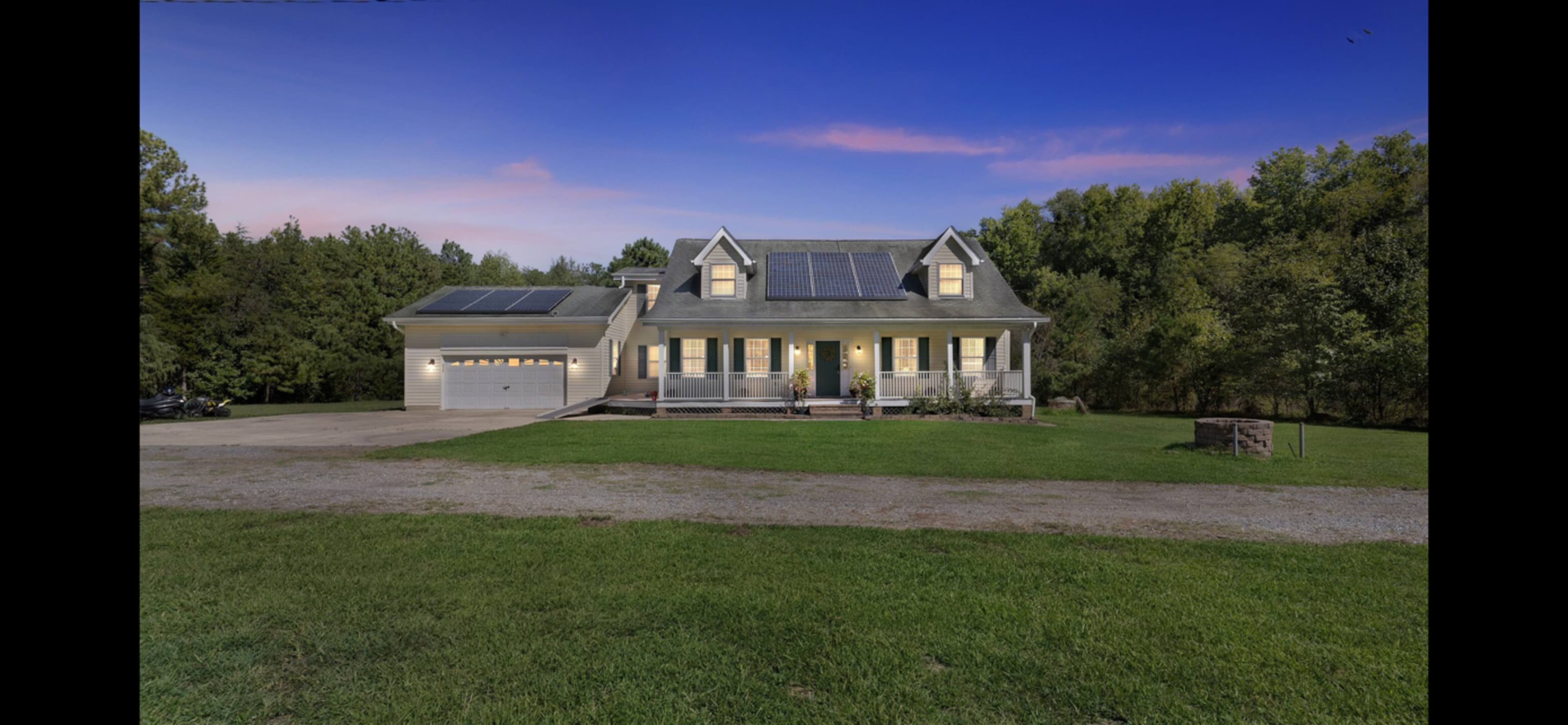 A house with solar panels on the roof, situated on a gravel driveway surrounded by trees at dusk.