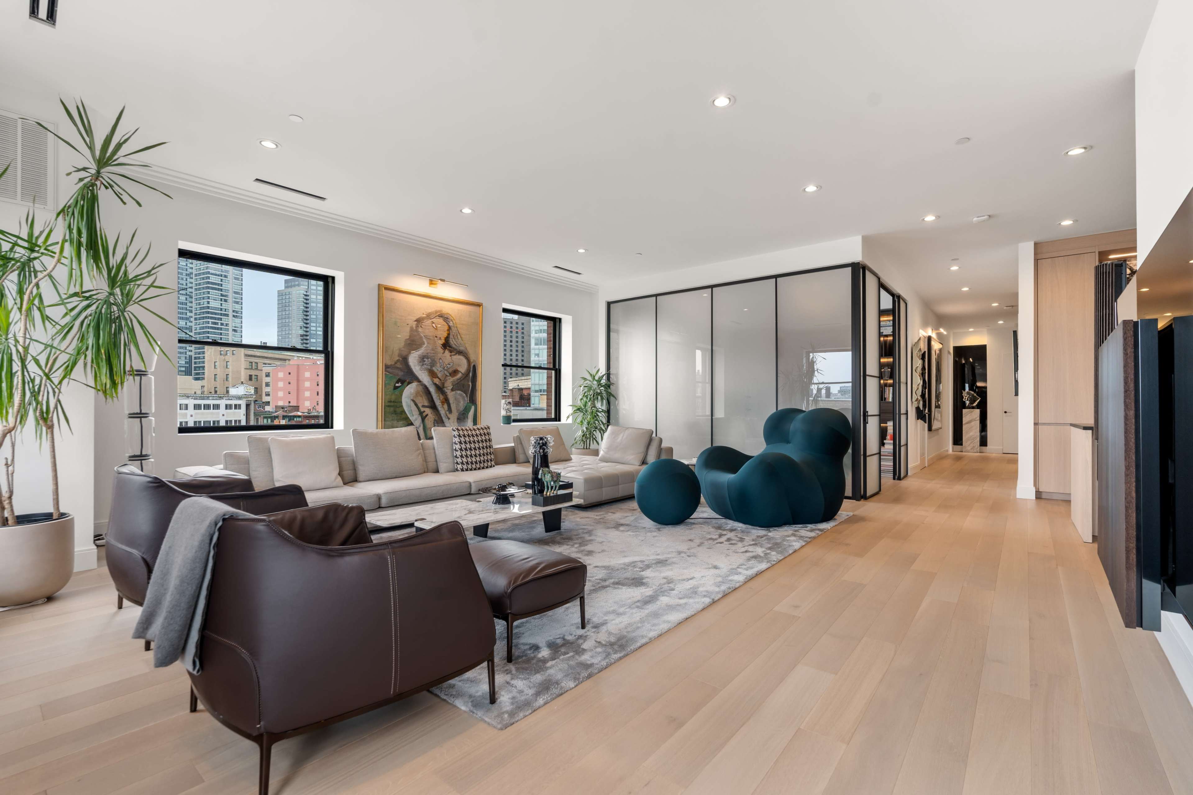 A modern living room with a large sectional sofa, a rug, and a glass-walled room in the background, featuring potted plants and artwork on the walls.