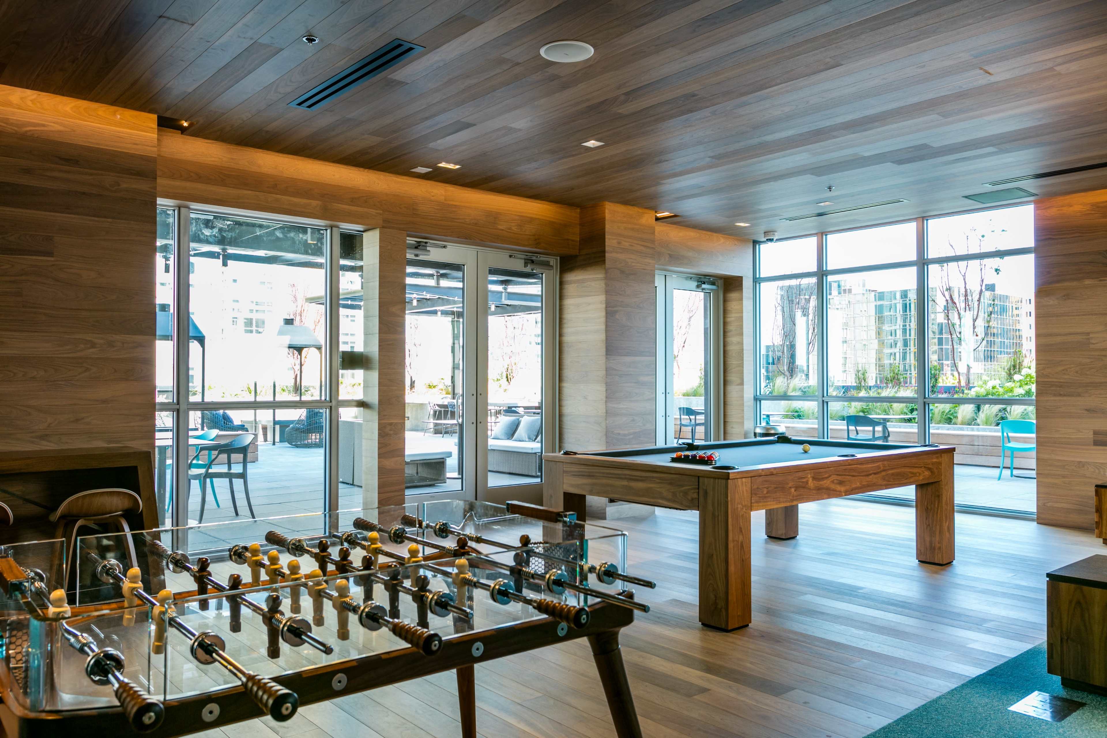 A modern game room with a foosball table and a billiard table, featuring large windows that provide natural light and views of an outdoor area.