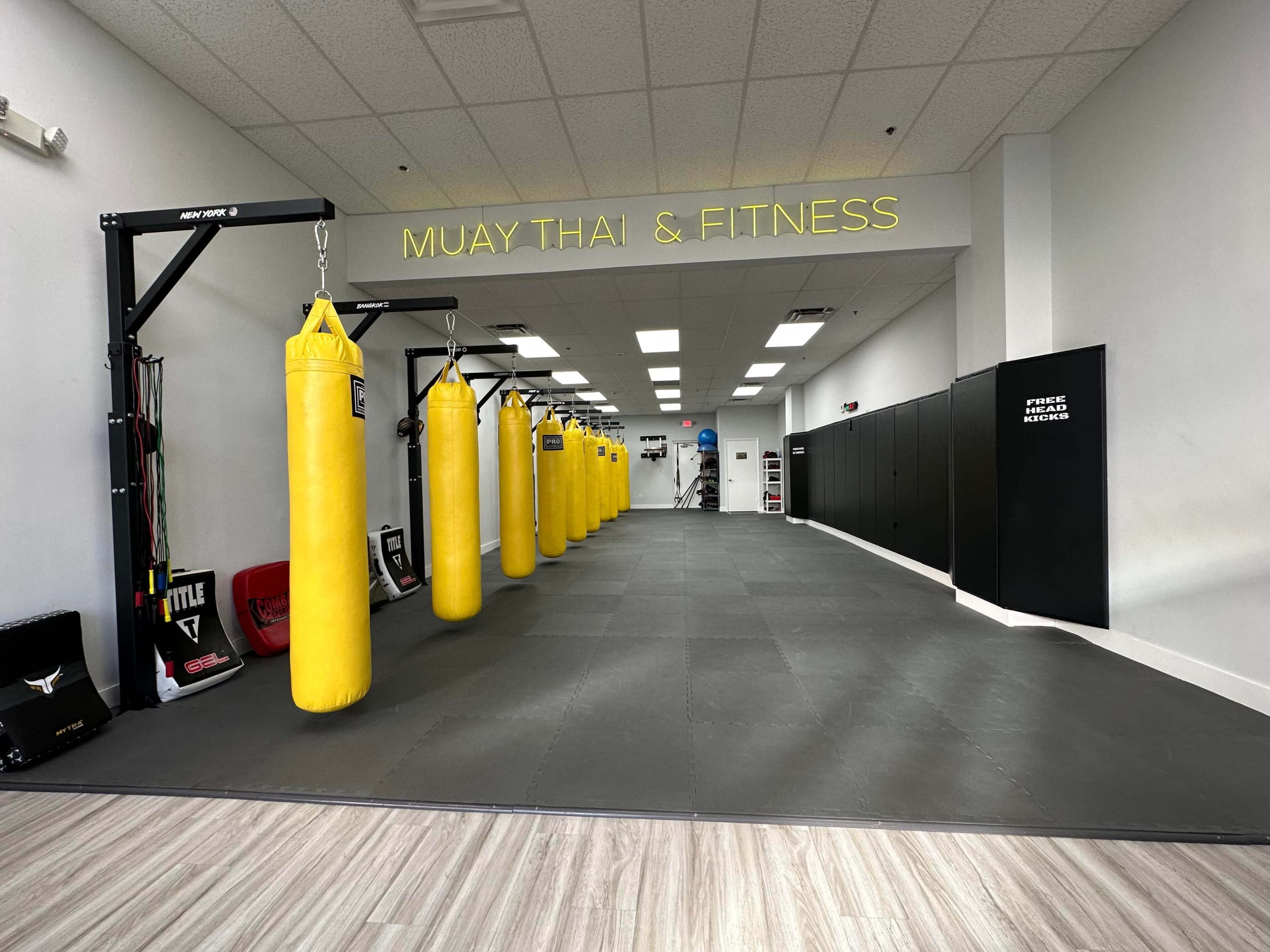 A Muay Thai and fitness gym featuring a row of yellow punching bags along one side of a spacious training area with black mats and padded walls.