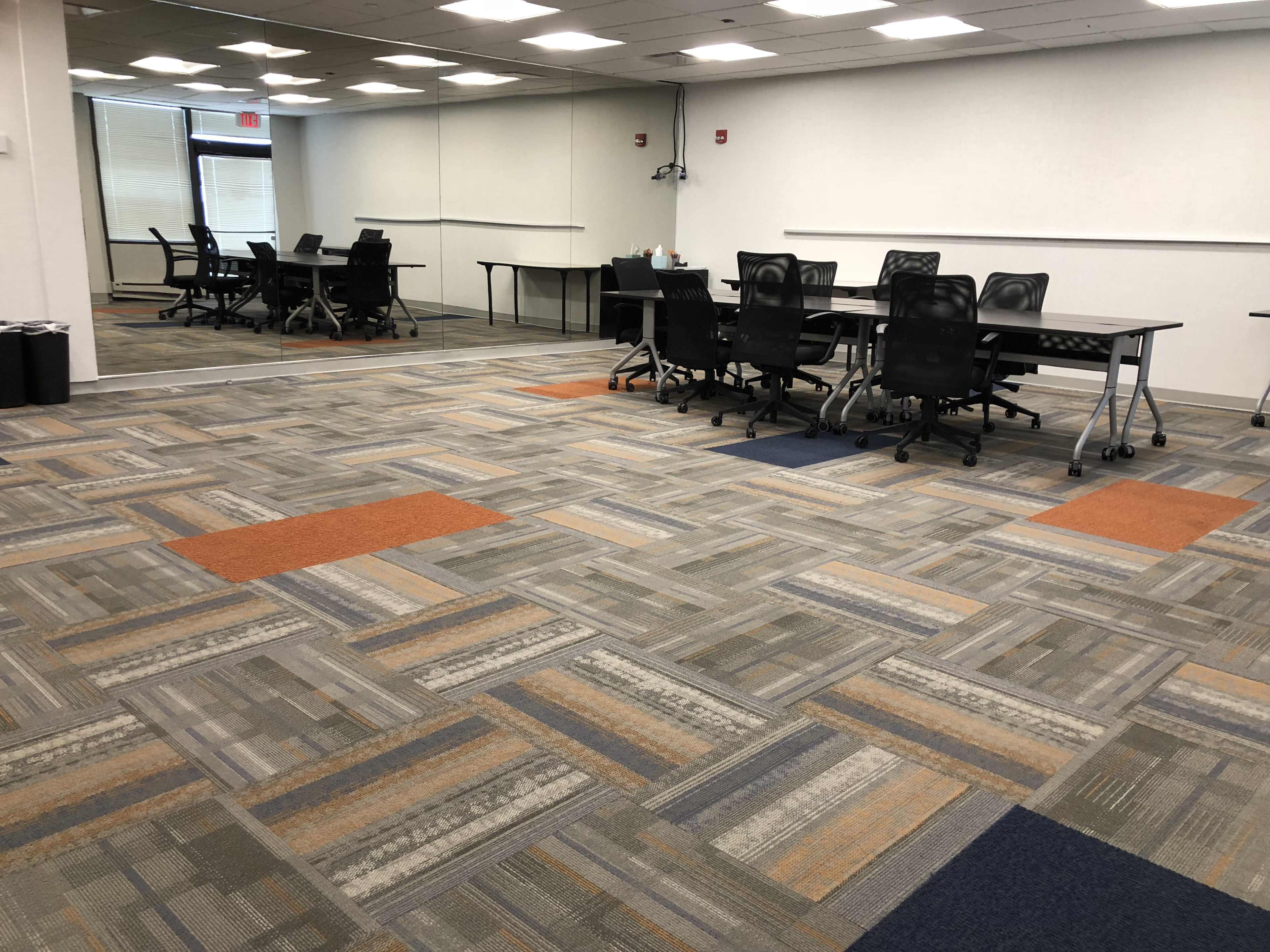 The image shows a modern meeting room with several black chairs around tables and a large mirrored wall, featuring patterned carpeting in shades of gray, orange, and blue.