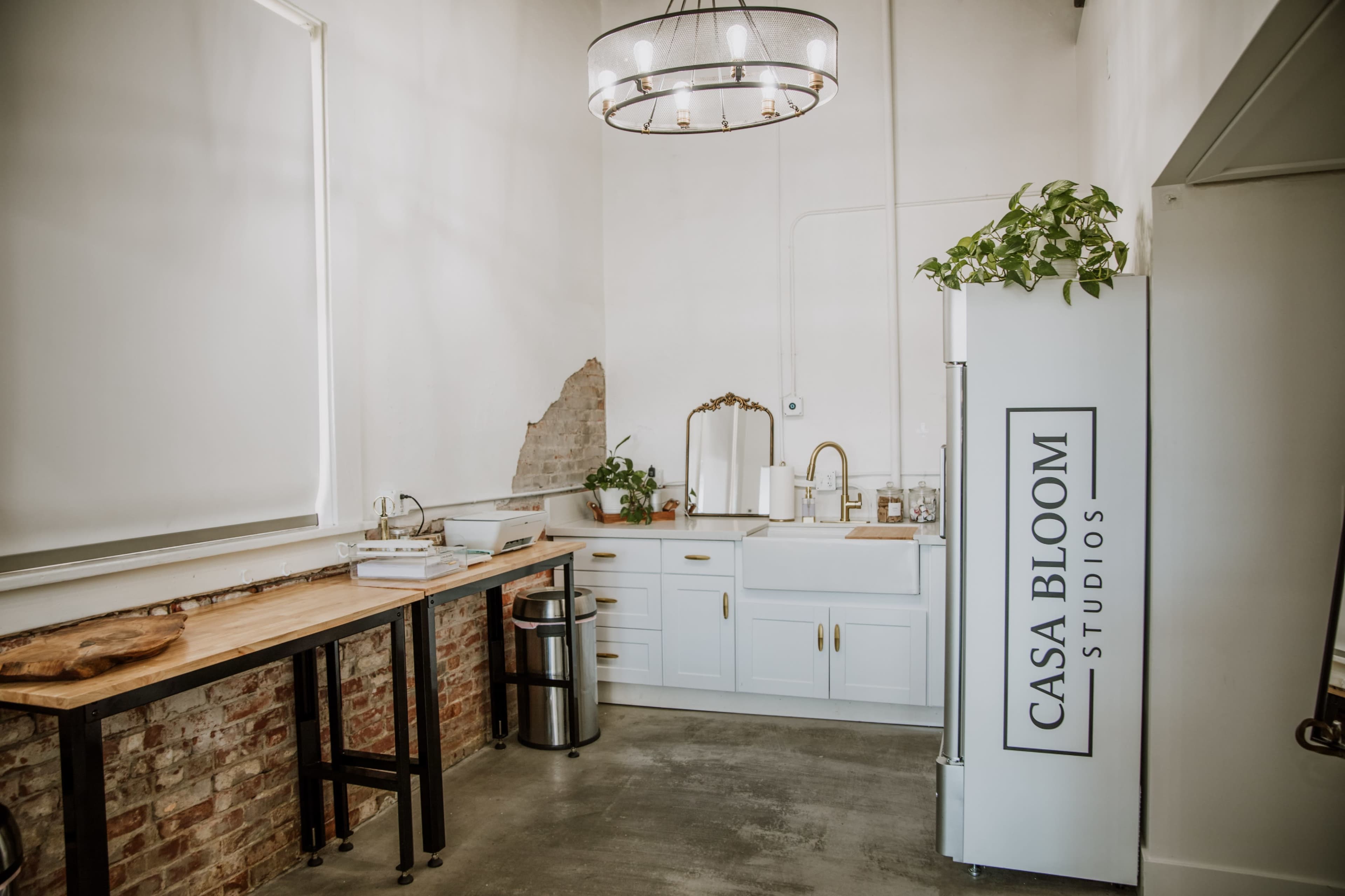 A bright studio kitchen area with a wooden table, modern cabinetry, and a refrigerator featuring the text "CASA BLOOM STUDIOS."
