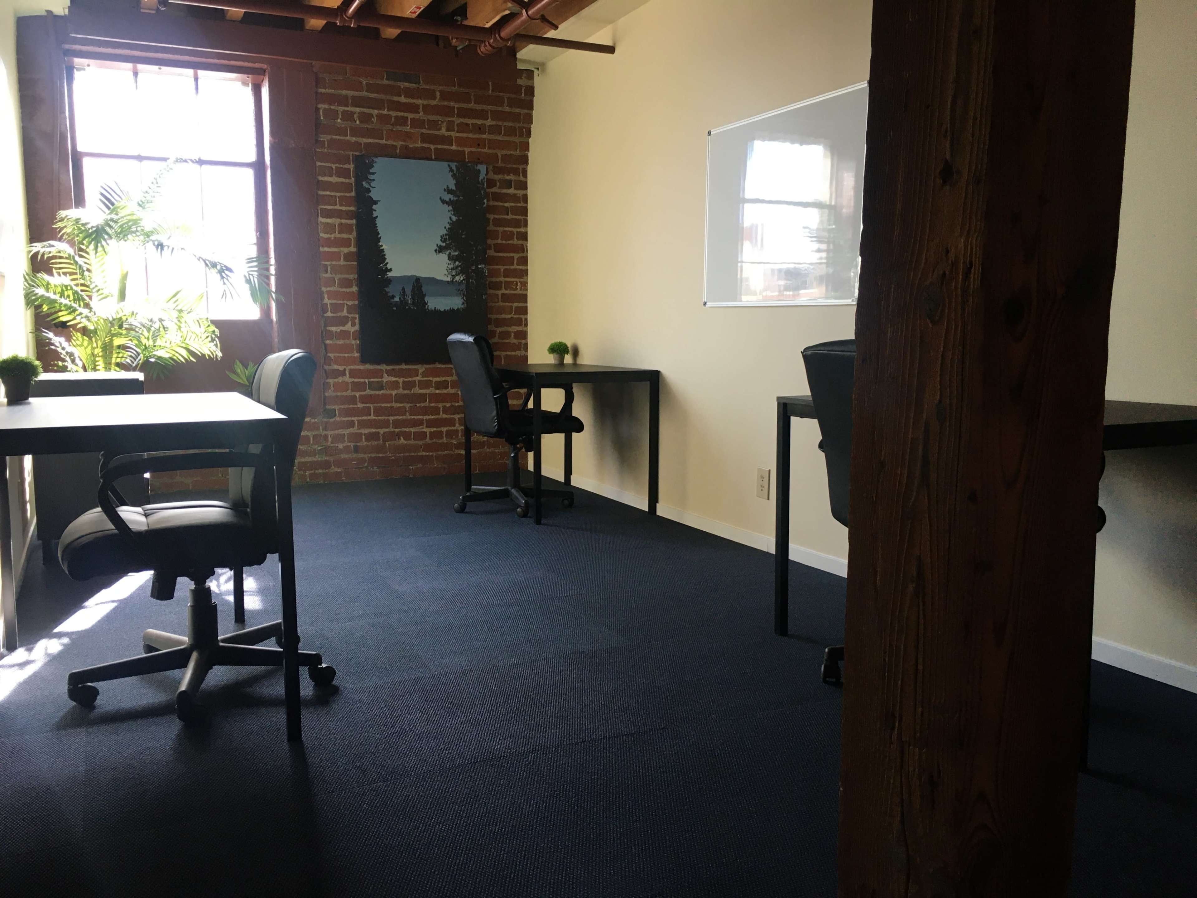 The image shows a minimalist office space with three black chairs at desks, a plant near a window, and a wall-mounted whiteboard.