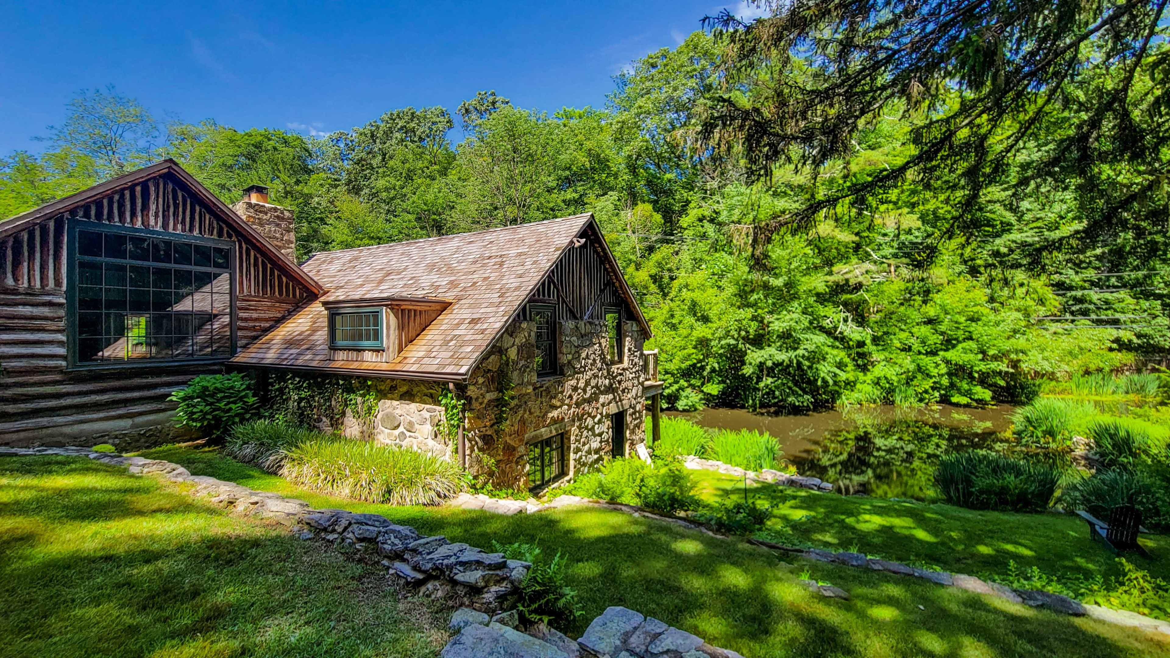 A rustic stone and wood house beside a serene pond, surrounded by lush greenery and trees.