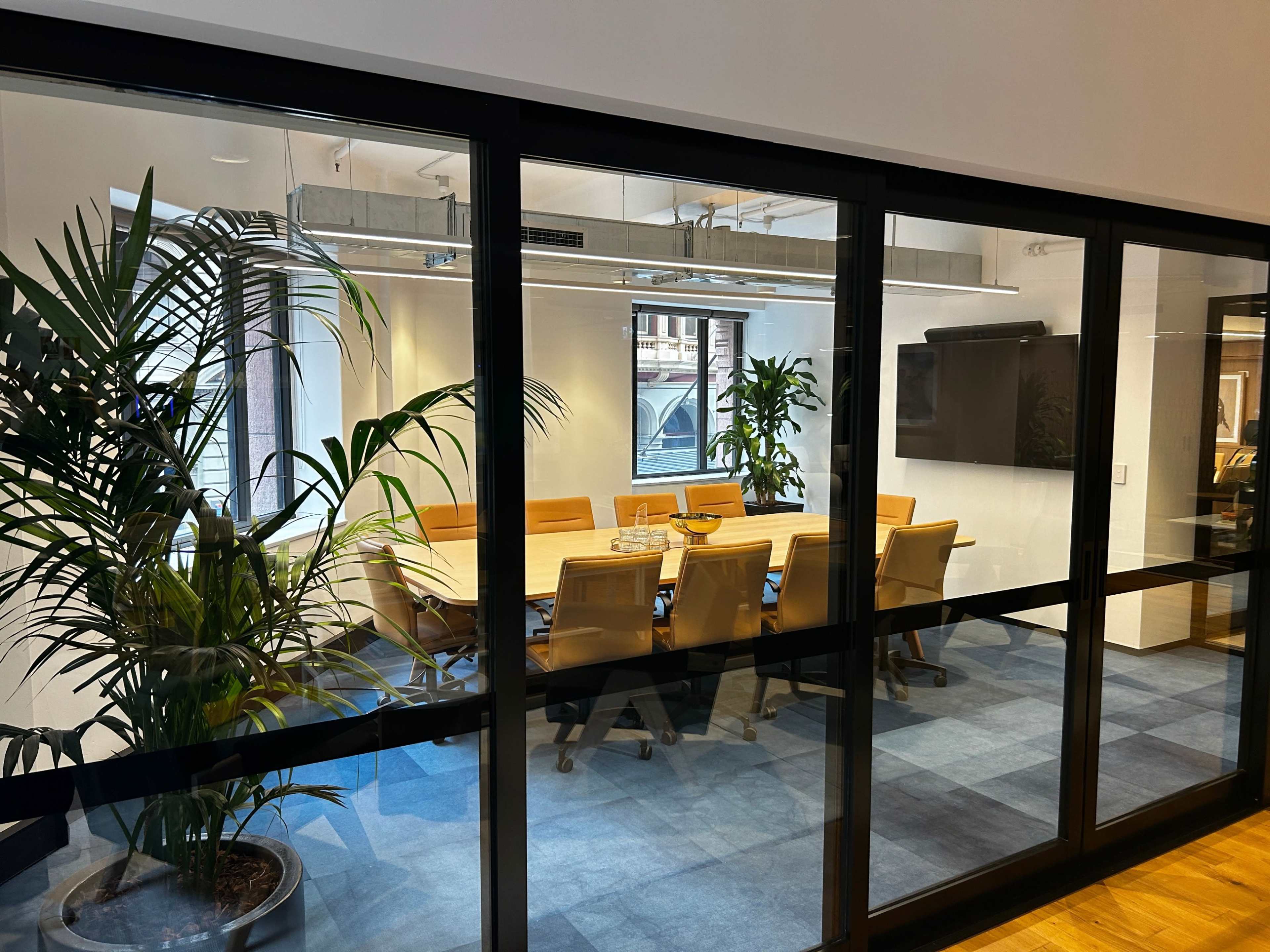 A glass-walled conference room features a large table surrounded by eight chairs and indoor plants, with a television mounted on the wall.