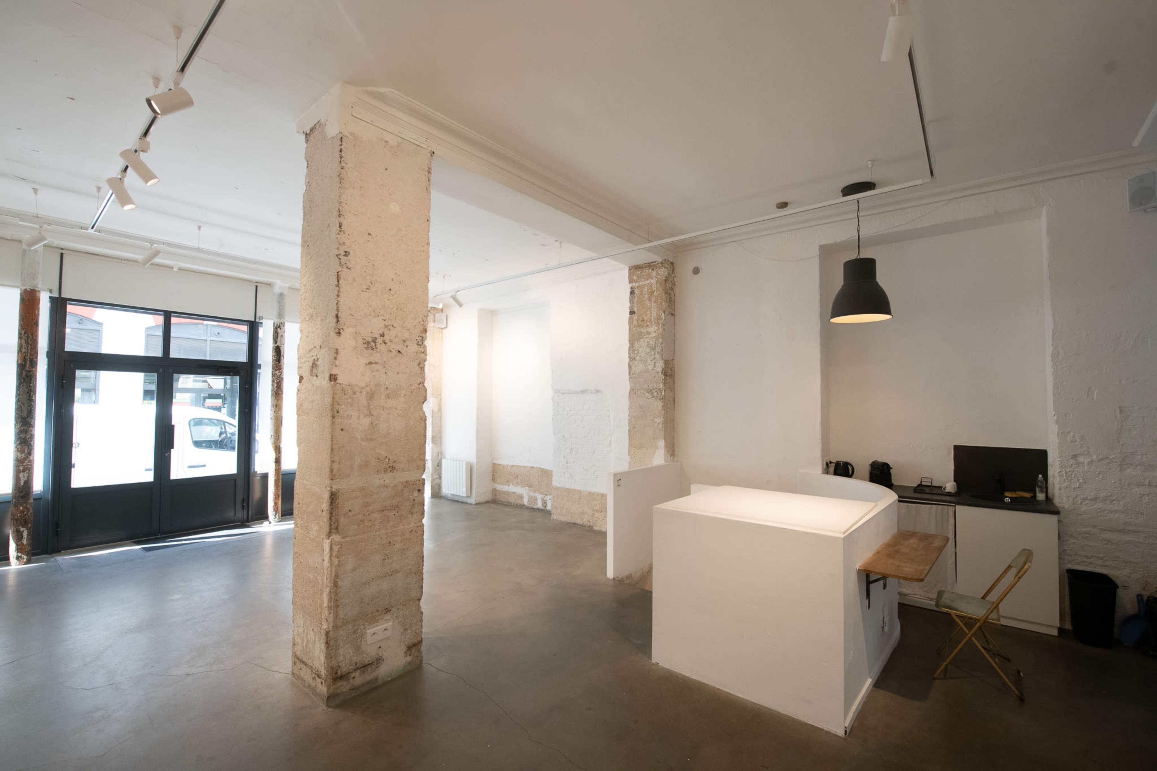 The image shows a minimalist interior space with a reception area, a desk with a chair, and large windows providing natural light.