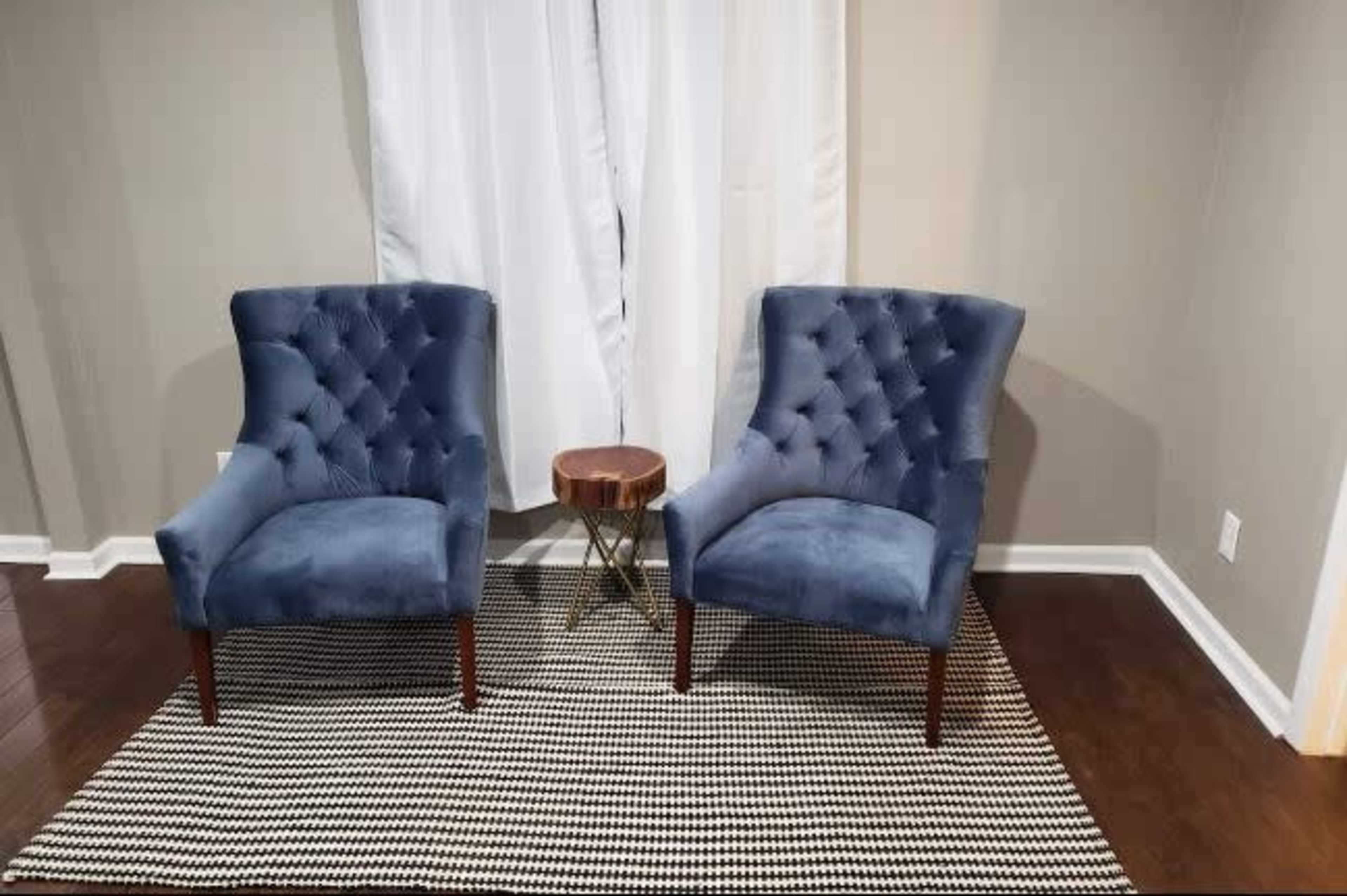 Two blue tufted chairs are positioned on a striped rug beside a small round table in a room with light-colored walls and a white curtain.
