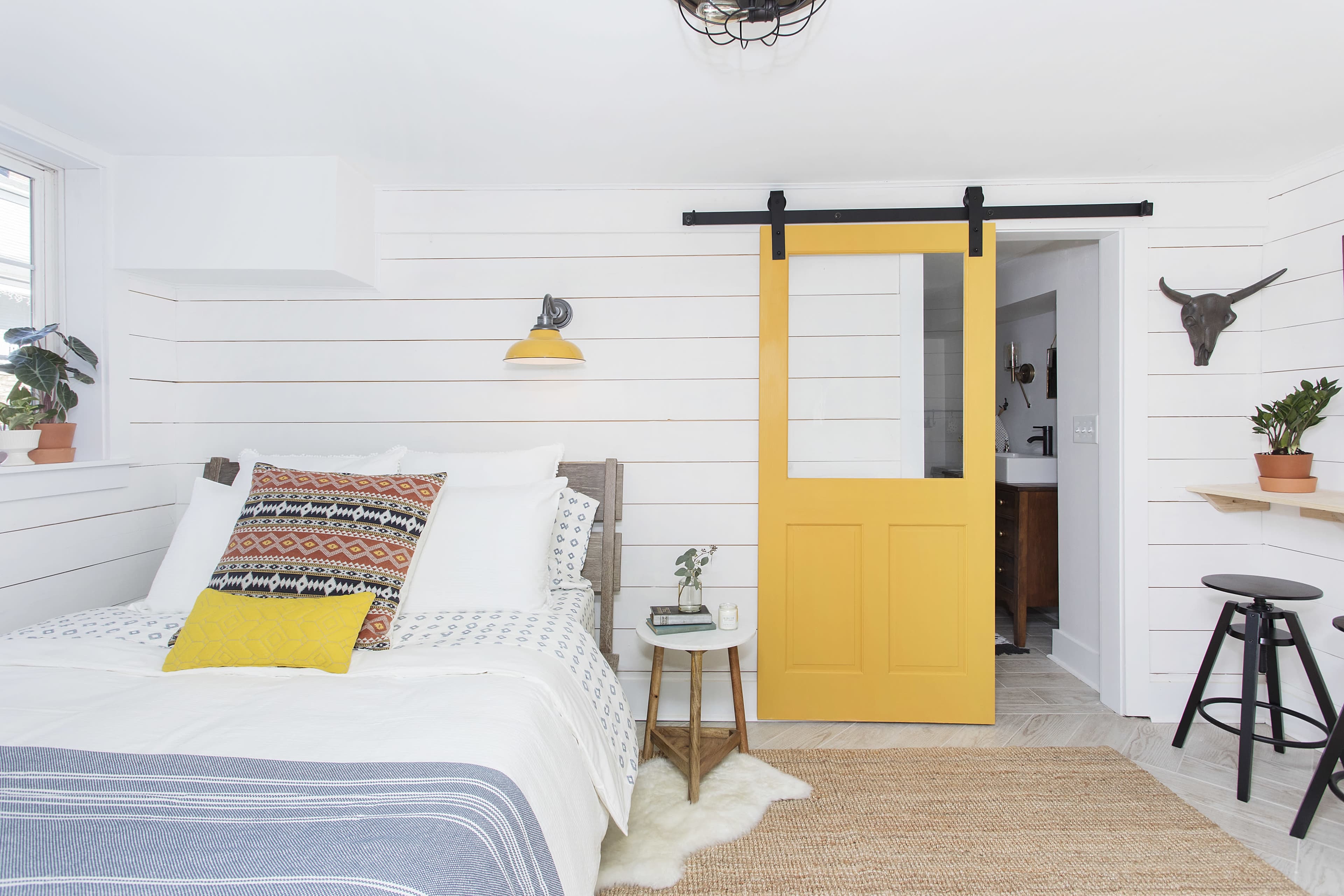 The scene depicts a modern bedroom featuring a yellow barn door, a bed with patterned linens, and wooden accents against white shiplap walls.