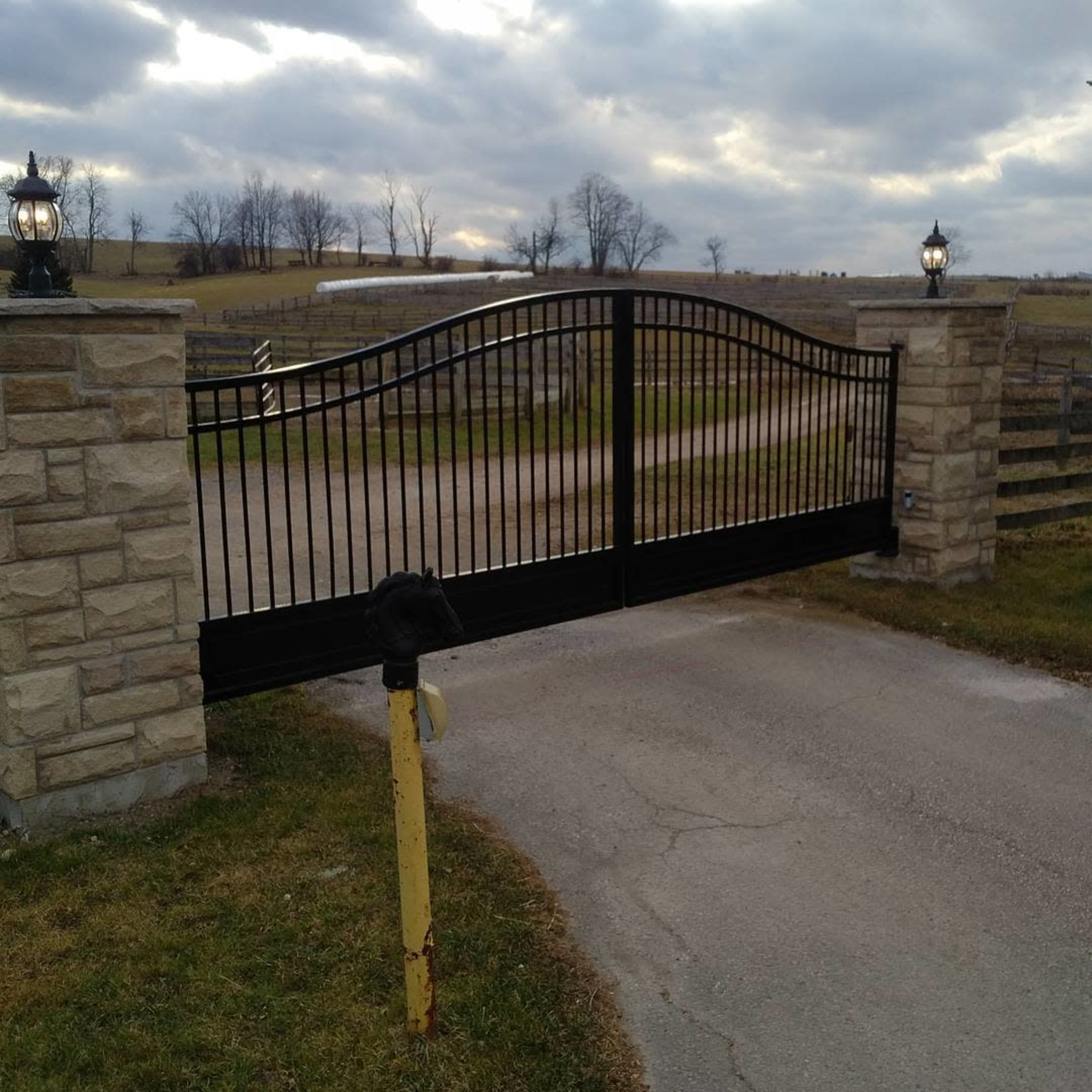 A black wrought iron gate is set between two stone pillars, leading into a gravel path surrounded by open fields and cloudy skies.