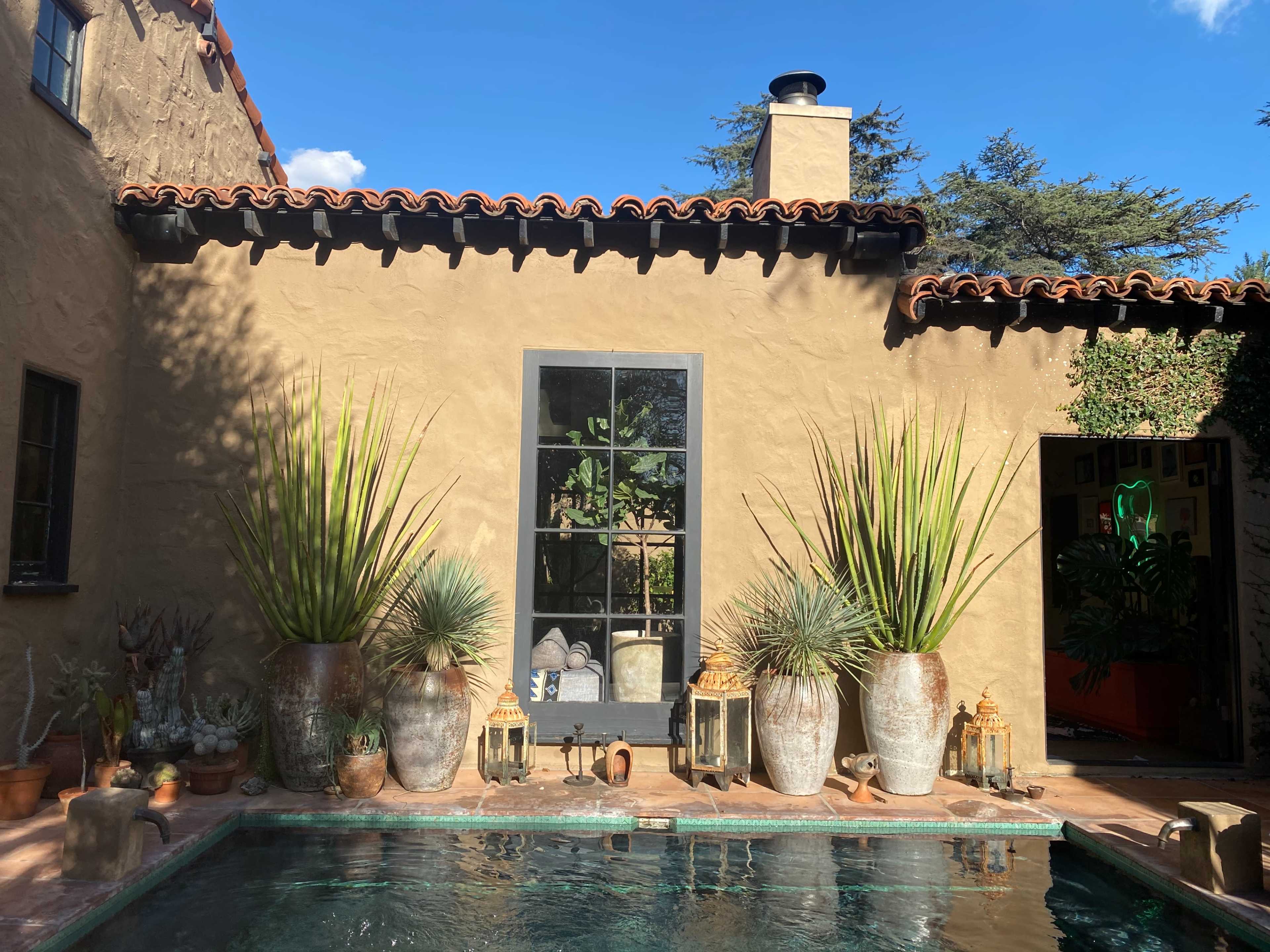 A courtyard with a pool, featuring tall potted plants and lanterns beside a textured wall and a large window.