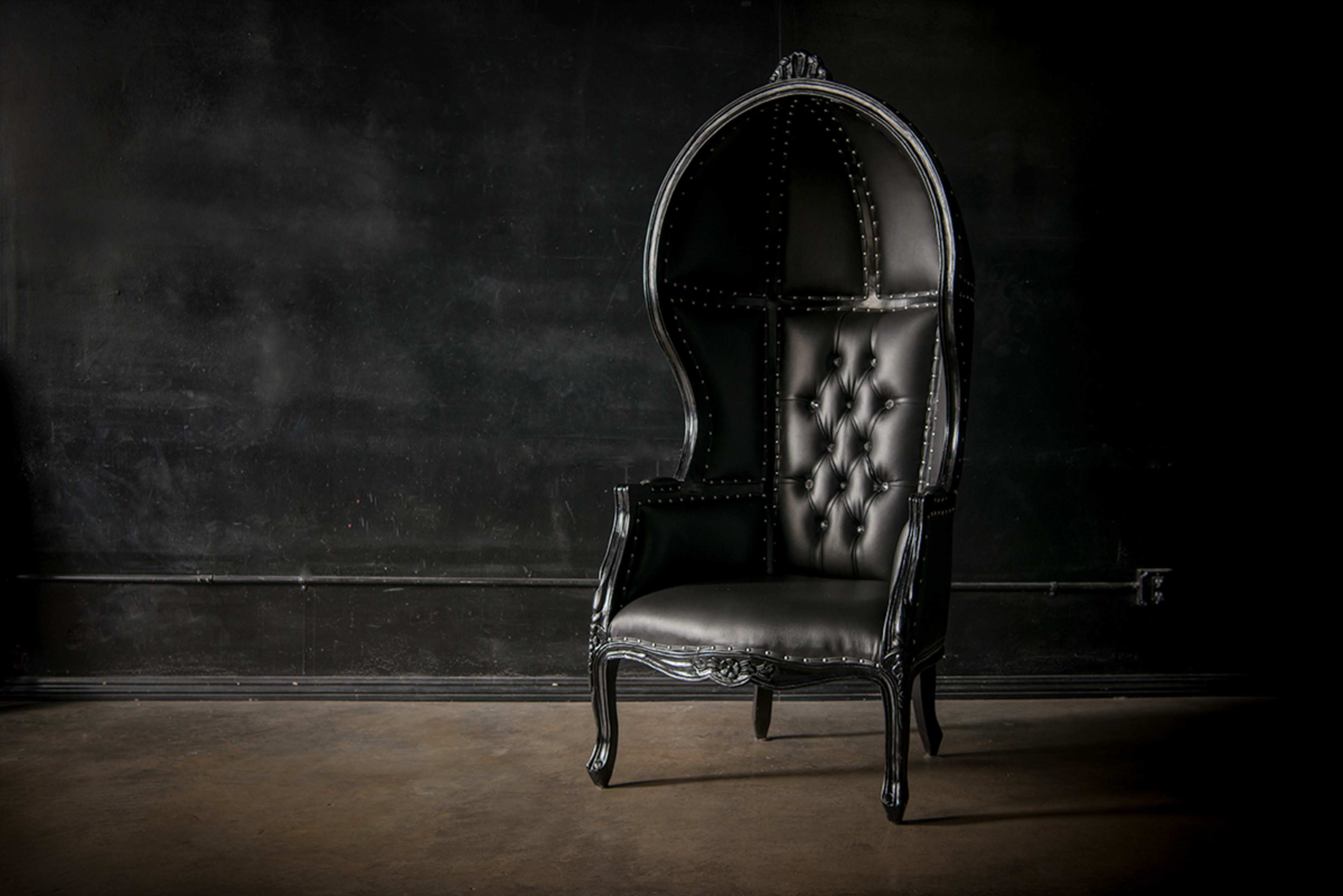 A large black throne chair with a high backrest and button-tufted upholstery stands against a dark wall.