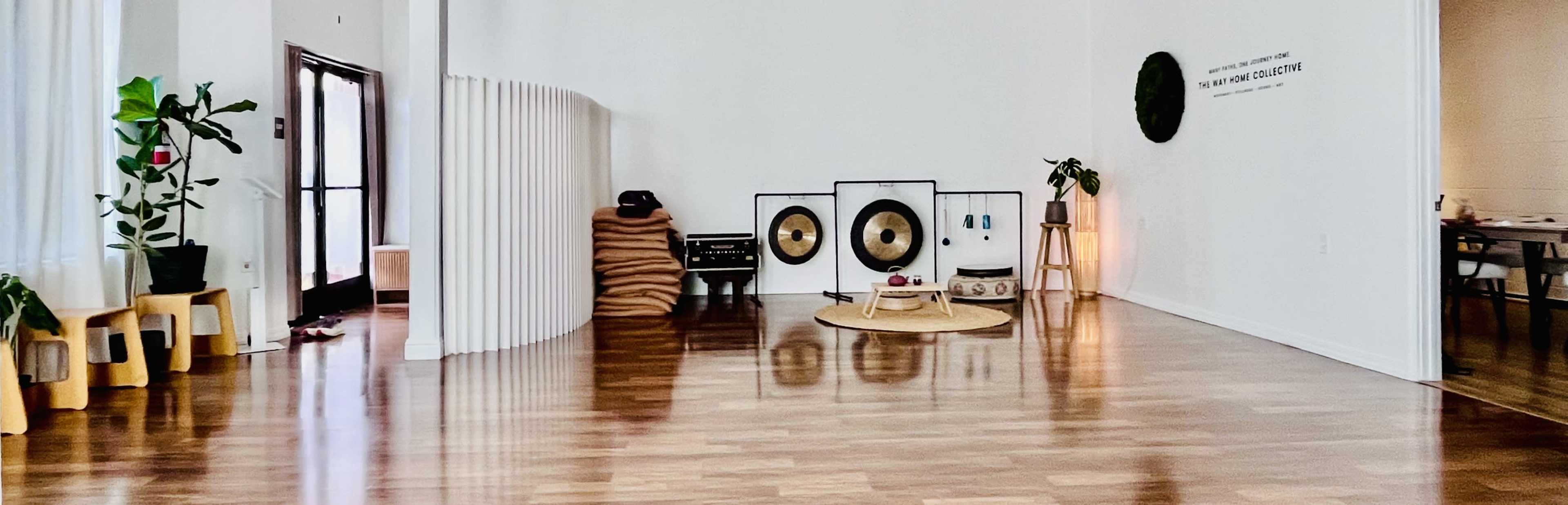 The image shows a spacious, minimalist interior with wooden flooring, featuring plants, musical instruments, and soft seating arrangements.