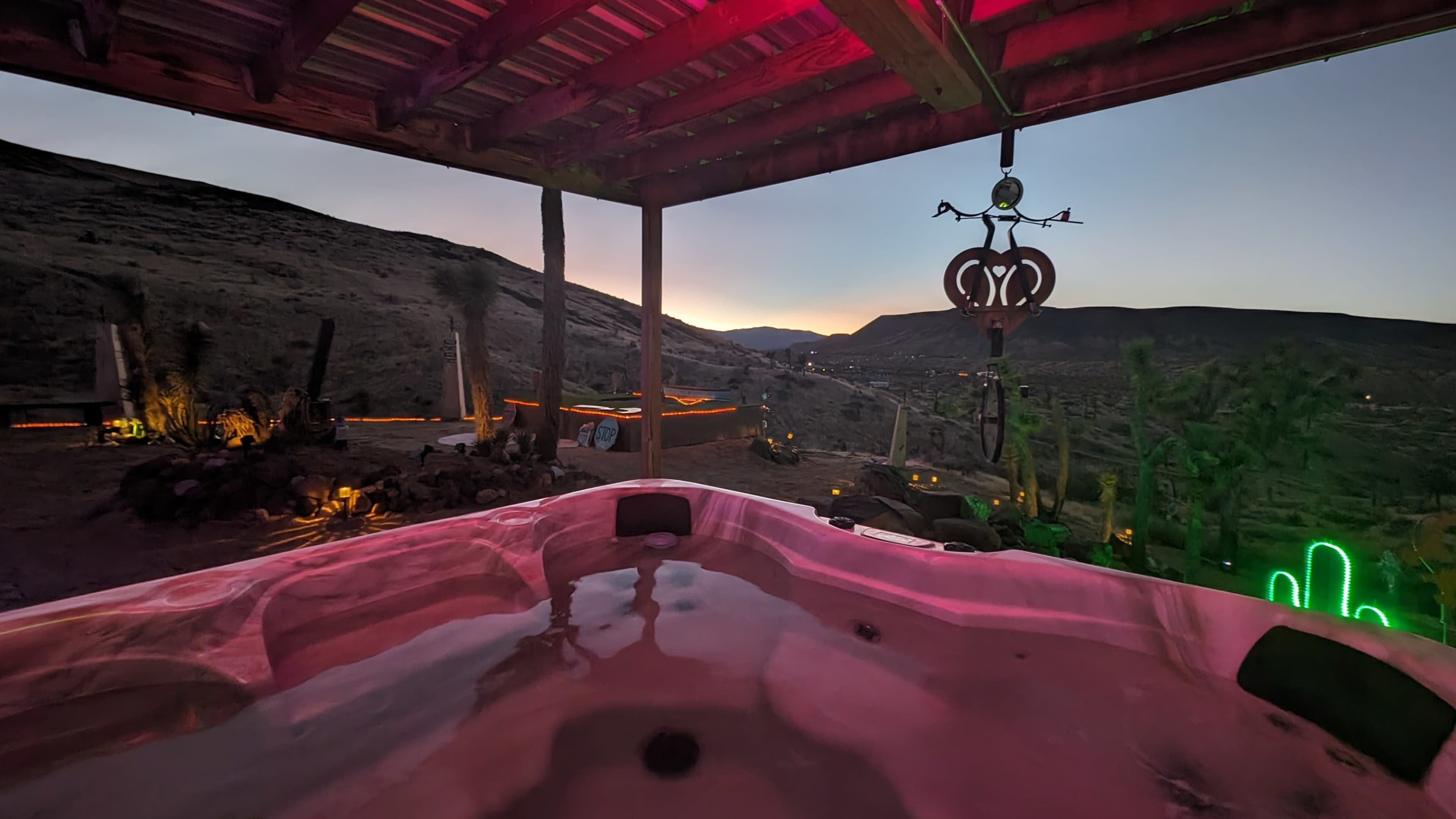The image shows a hot tub on a patio with colorful lights, overlooking a mountainous landscape at dusk.