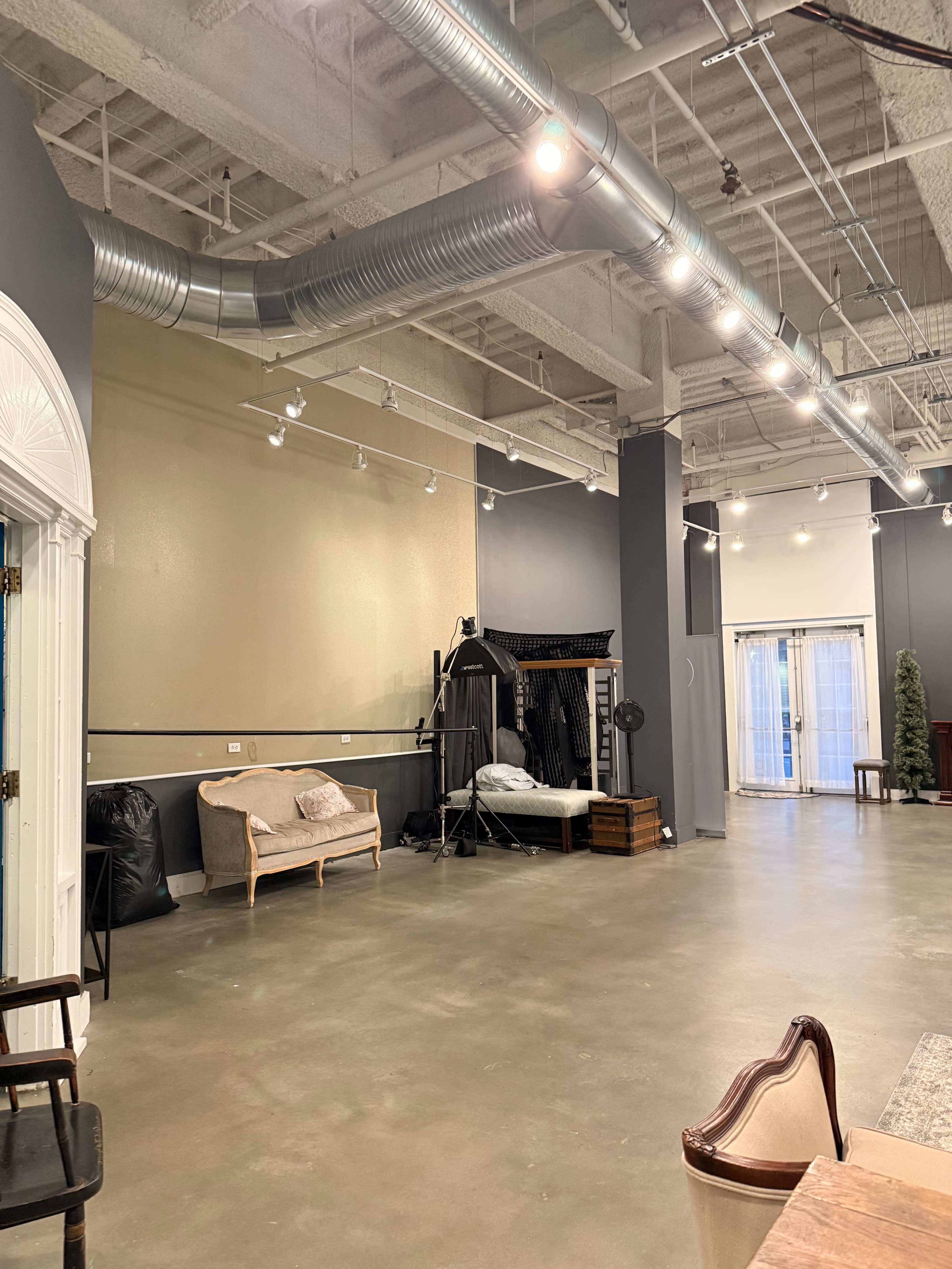 The image shows an empty industrial-style indoor space with gray walls, polished concrete flooring, and various seating options, including a vintage couch and a coat rack.