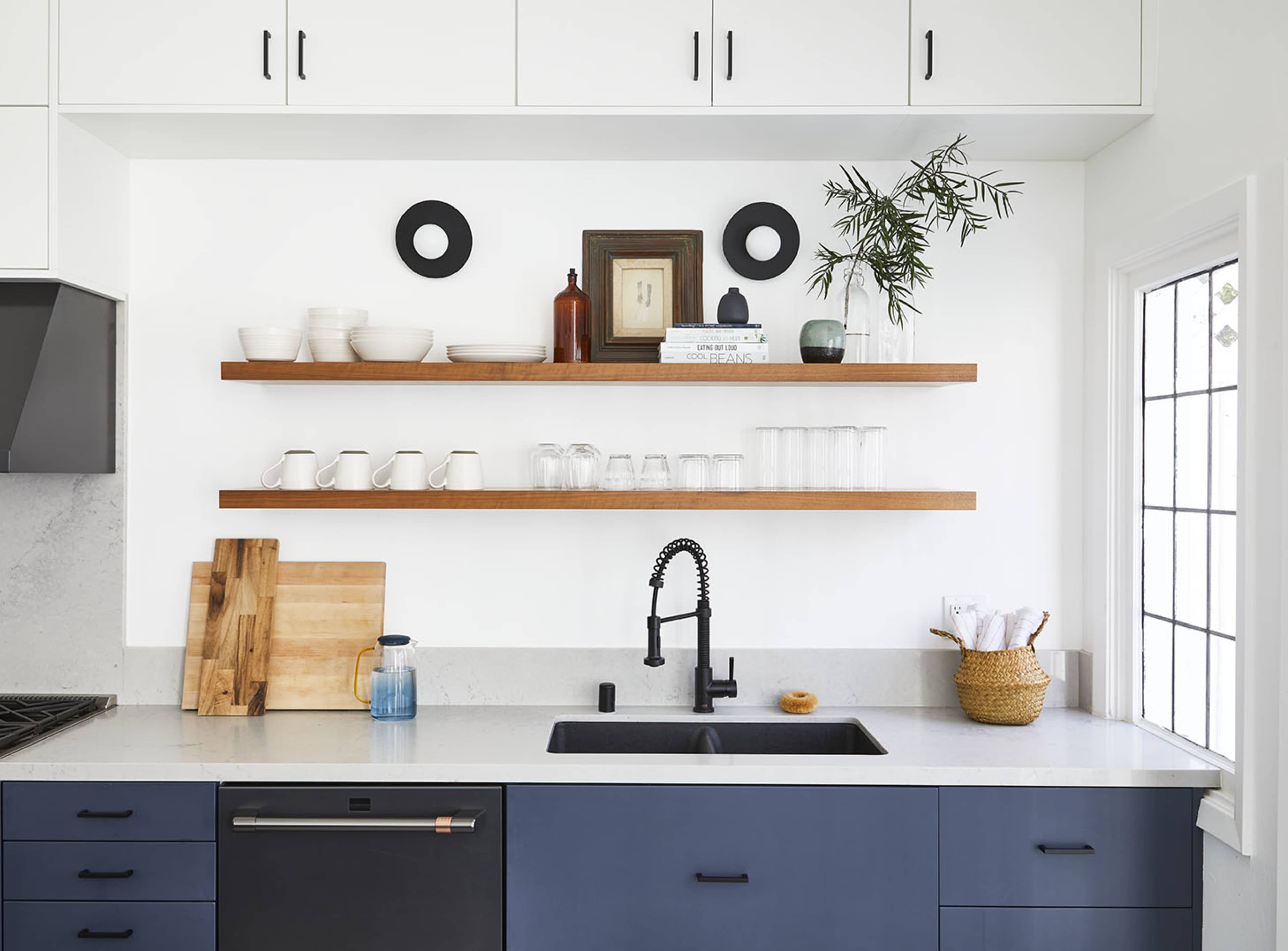 A modern kitchen features a black sink, wooden shelves with dishware, and a small plant, against a white wall and light wood cabinetry.